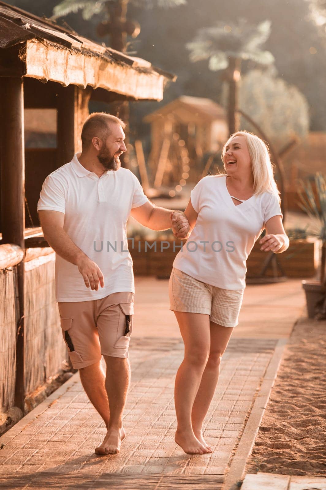 A happy married couple in shorts and T-shirts walks down the street at sunset in summer by Lobachad