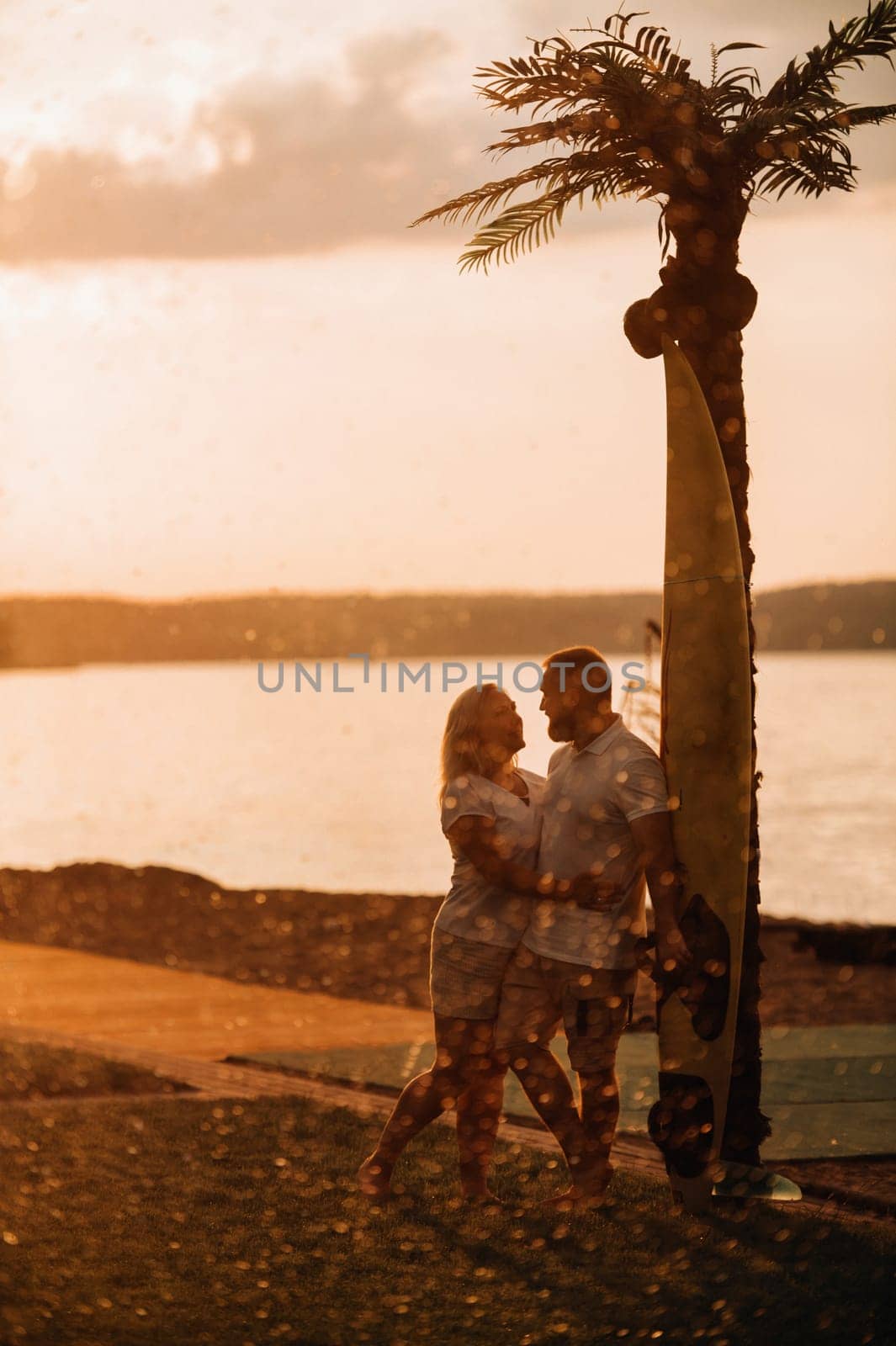 A happy married couple in shorts and T-shirts hug under a palm tree at sunset in summer by Lobachad