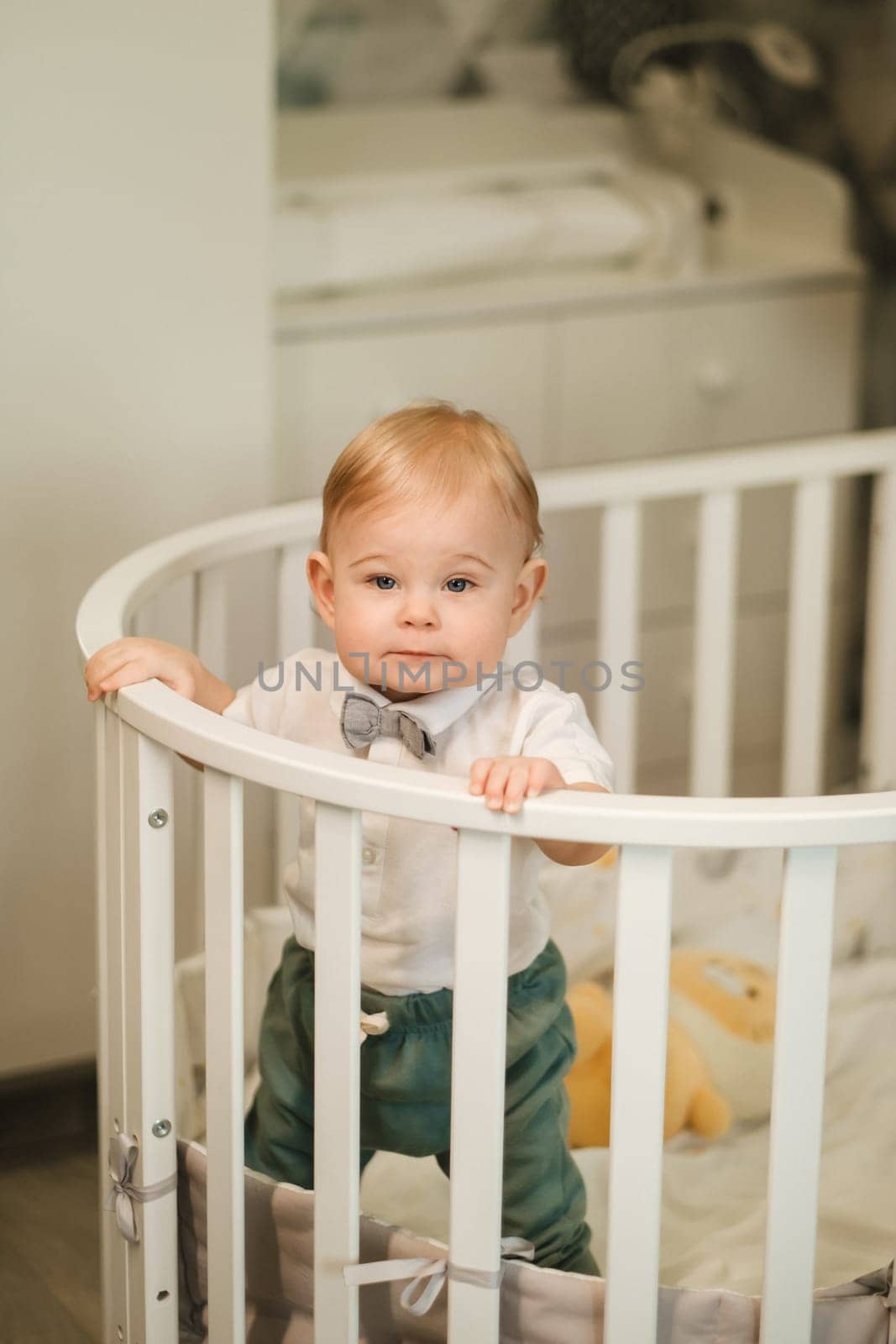 Portrait of a cheerful little boy in a white shirt with a bow tie standing in a crib.