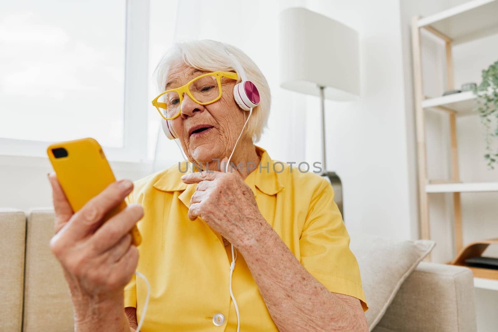 Happy elderly woman watching video call on phone and listening to music on headphones, surprise and open mouth, bright modern interior, lifestyle online communication. High quality photo