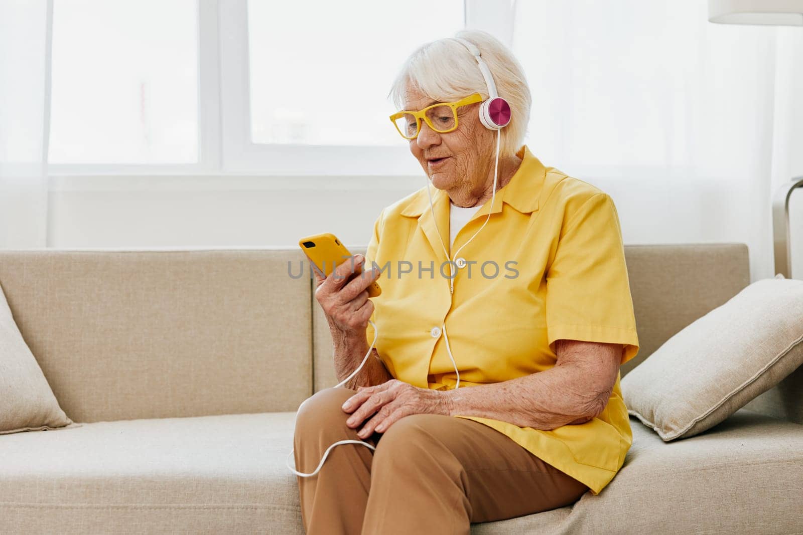 Happy elderly woman looking into her phone video call with headphones smile, technology for communication, bright modern interior, lifestyle online communication. High quality photo