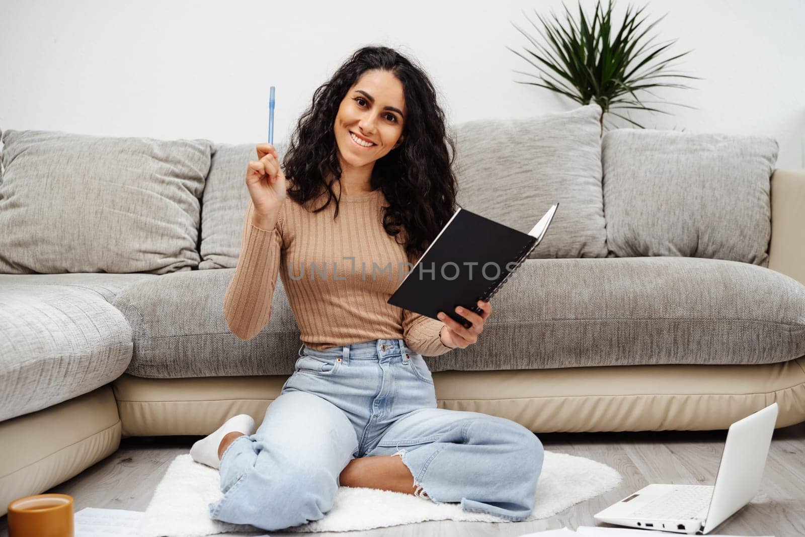 Young beautiful Arab woman holds a notebook in her hand and makes a wish list. She took a pen and is ready to paint everything point by point. The girl sits on the floor and smiles brightly at home.