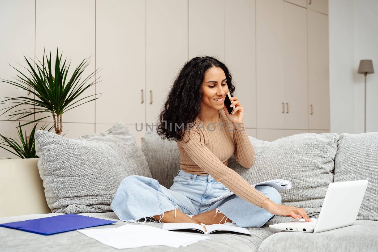 Young beautiful woman of Arab works remotely at home. One hand is typing on the laptop while the other hand is holding the phone. The girl joyfully communicates on the phone about the tasks.