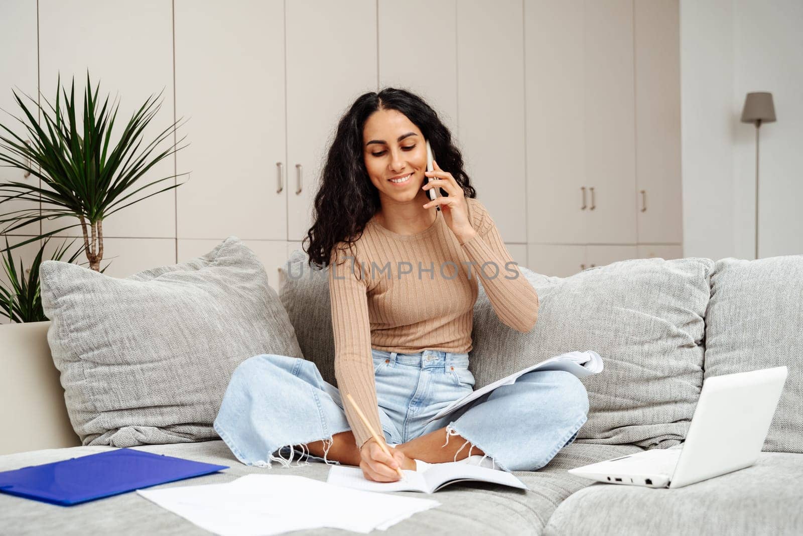 Young beautiful female student of Arab appearance sits on the sofa at home and studies. She holds a phone in one hand and takes notes in the other. Carries on a pleasant conversation about the subject of the task.