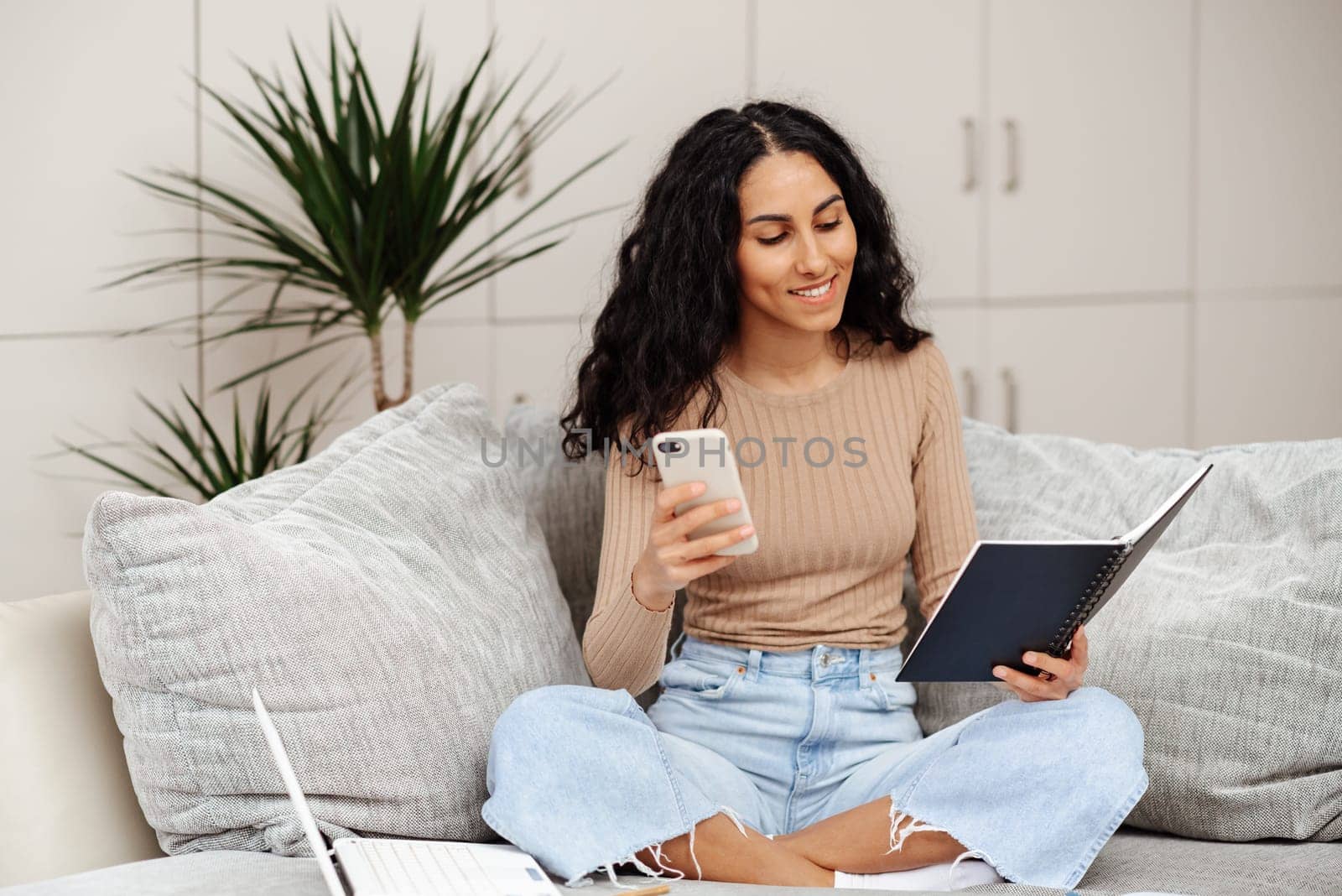 Shot of pretty young woman using her mobile phone while working with laptop sitting on sofa at home. Chooses a course of study checks his notes