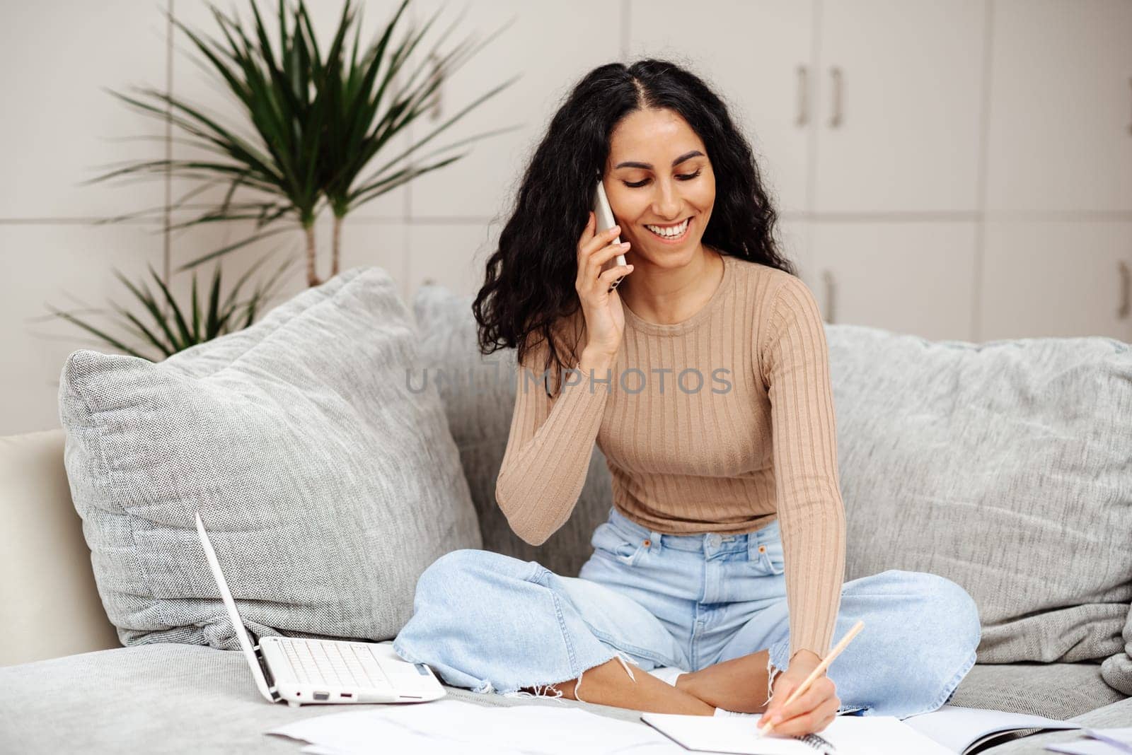 Education concept. Happy middle eastern woman making call about task, studying at home with laptop, taking notes while watching webinar or lesson. Writes homework in a notebook sitting on the sofa