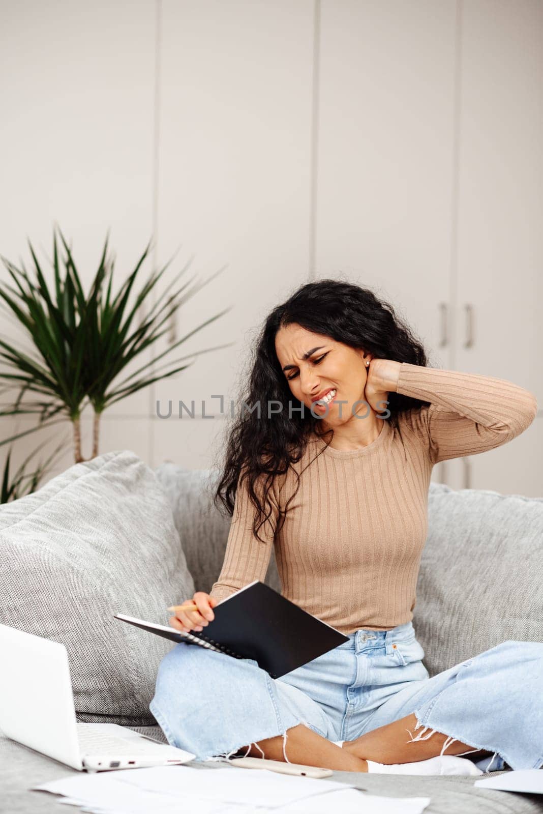 Frowning Arab woman sitting on the sofa near the computer, break, touching the neck, suffering from soreness, pain caused by poor bad posture, sedentary work, sedentary work, sitting at the laptop for a long period of concept image