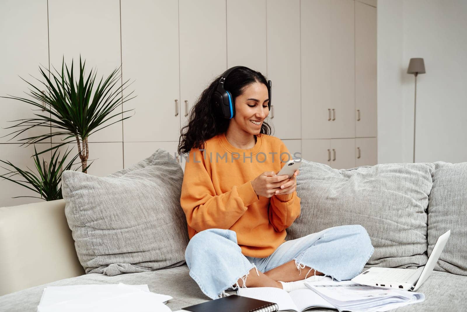 Arab teenager girl looks through messages in the phone and writes down homework. Listening to the seminar through headphones. The concept of distance learning. Female student happily studying at home
