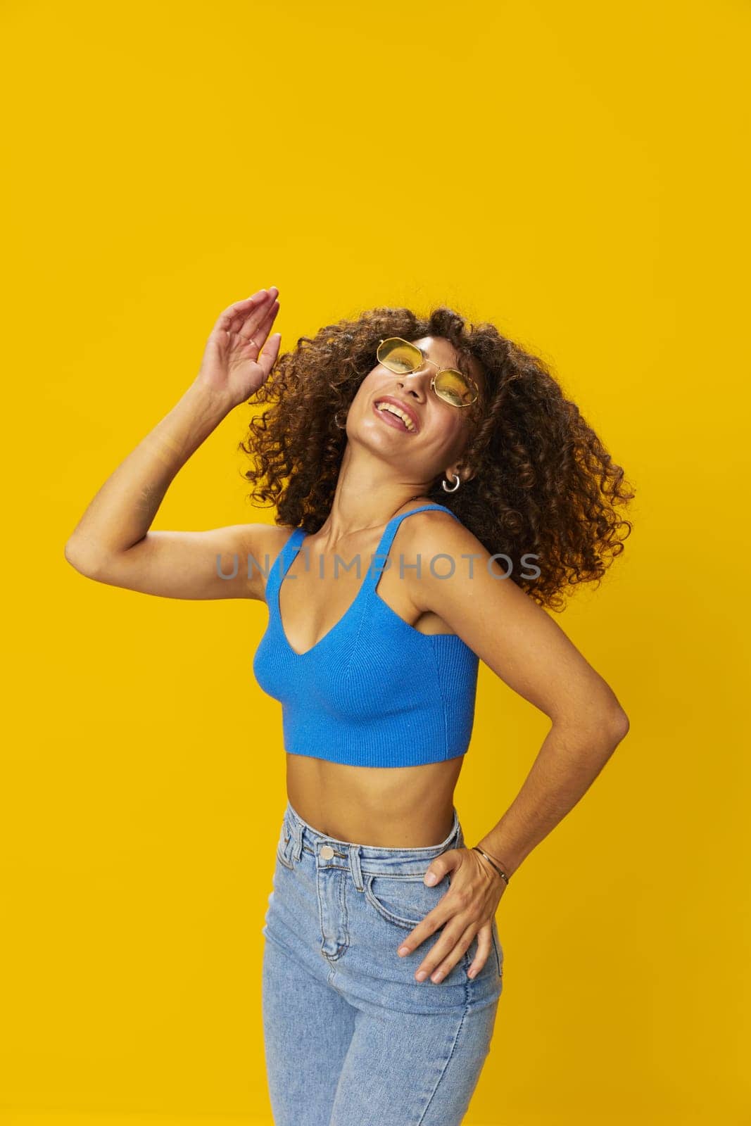 Woman with curly afro hair in a blue T-shirt on a yellow background dancing flying hair with sunglasses yellow, hand signs, look into the camera, smile with teeth and happiness, copy space by SHOTPRIME
