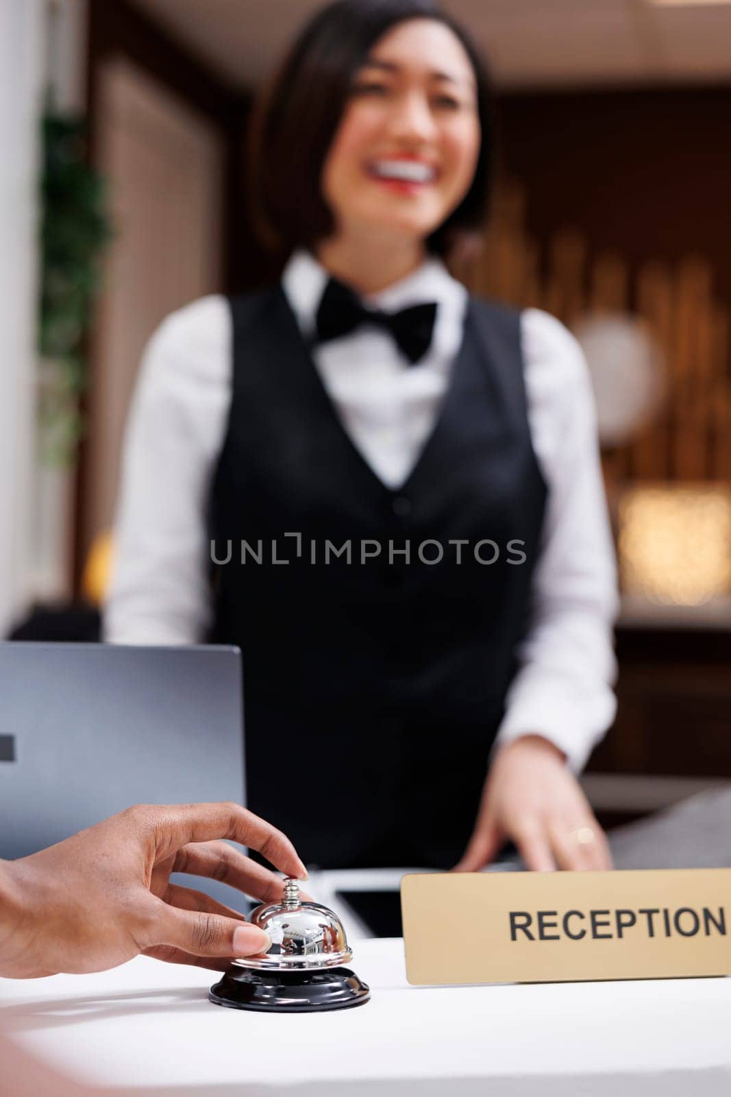 Person ringing bell at hotel reception with front desk staff working on check in and registration process. Young man using service bell in resort lobby, receptionist welcoming people. Close up.