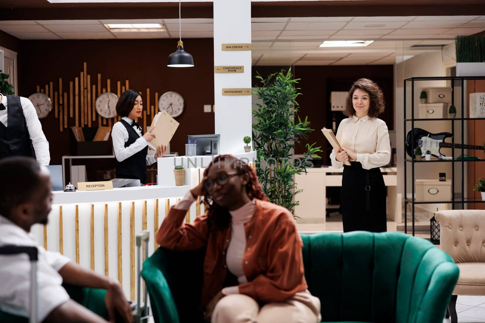 Hotel manager approaching guests on sofa, preparing to talk to people about accommodation and luxury resort service. Administrator standing in reception lobby, talking to guests.