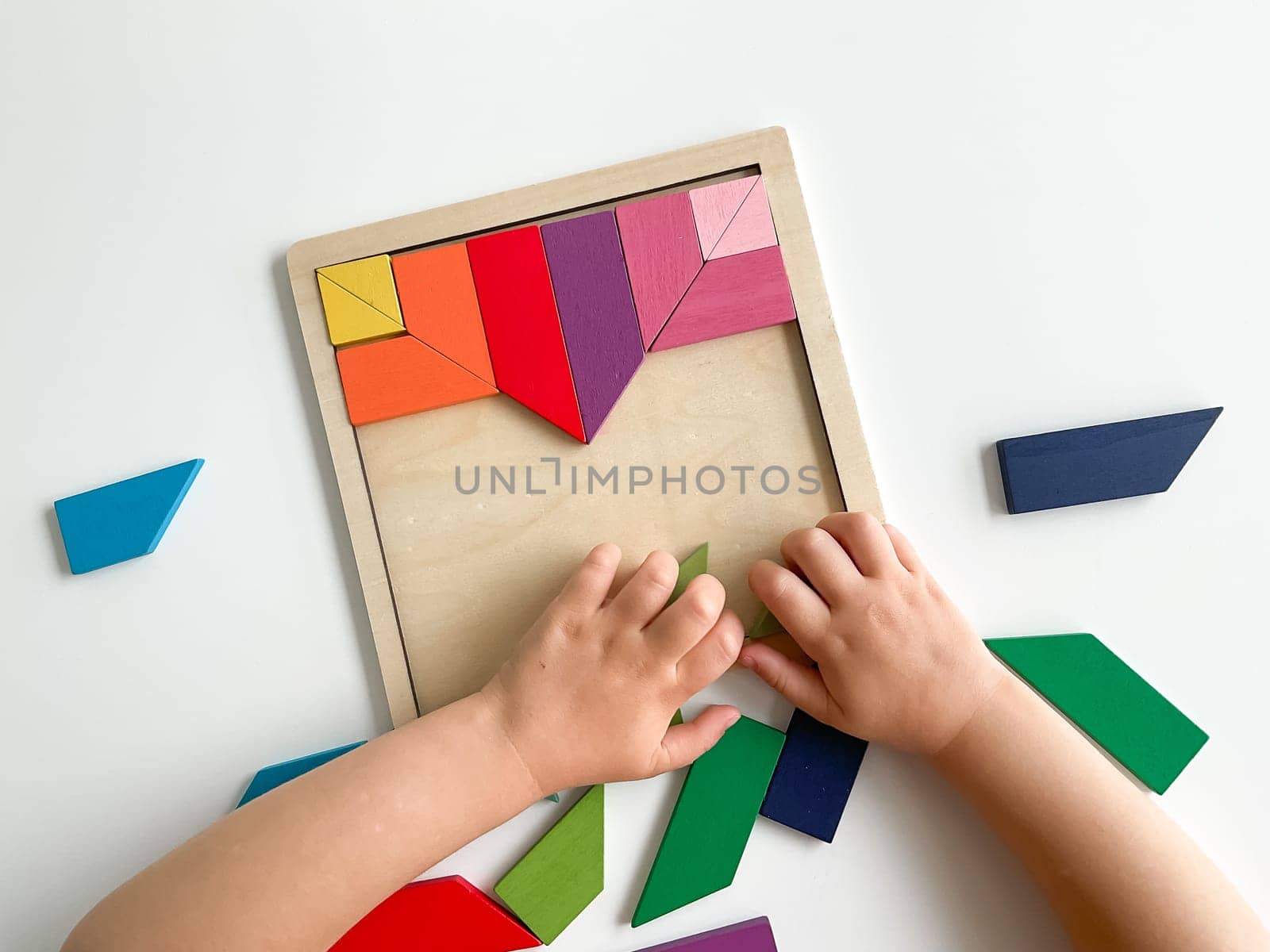 childs hand collects multicolored wooden mosaic on white background. child solves colorful tangram by Lunnica