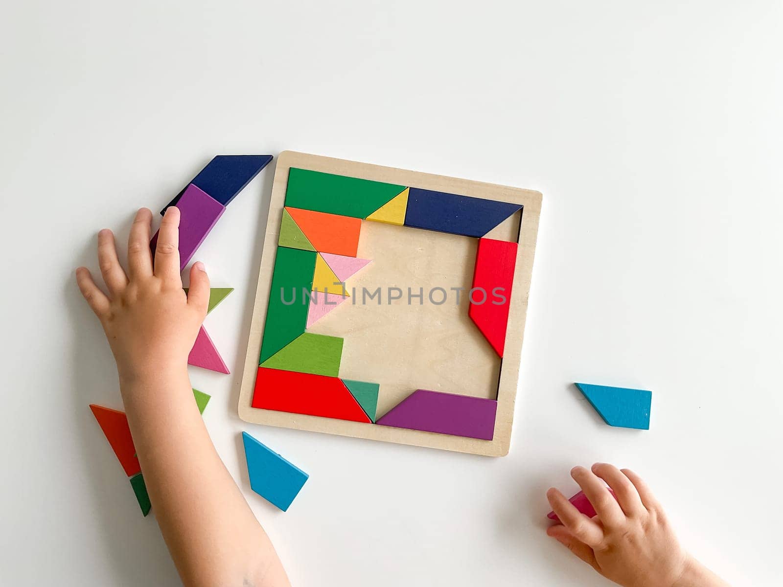 childs hand collects multicolored wooden mosaic on white background. child solves colorful tangram by Lunnica