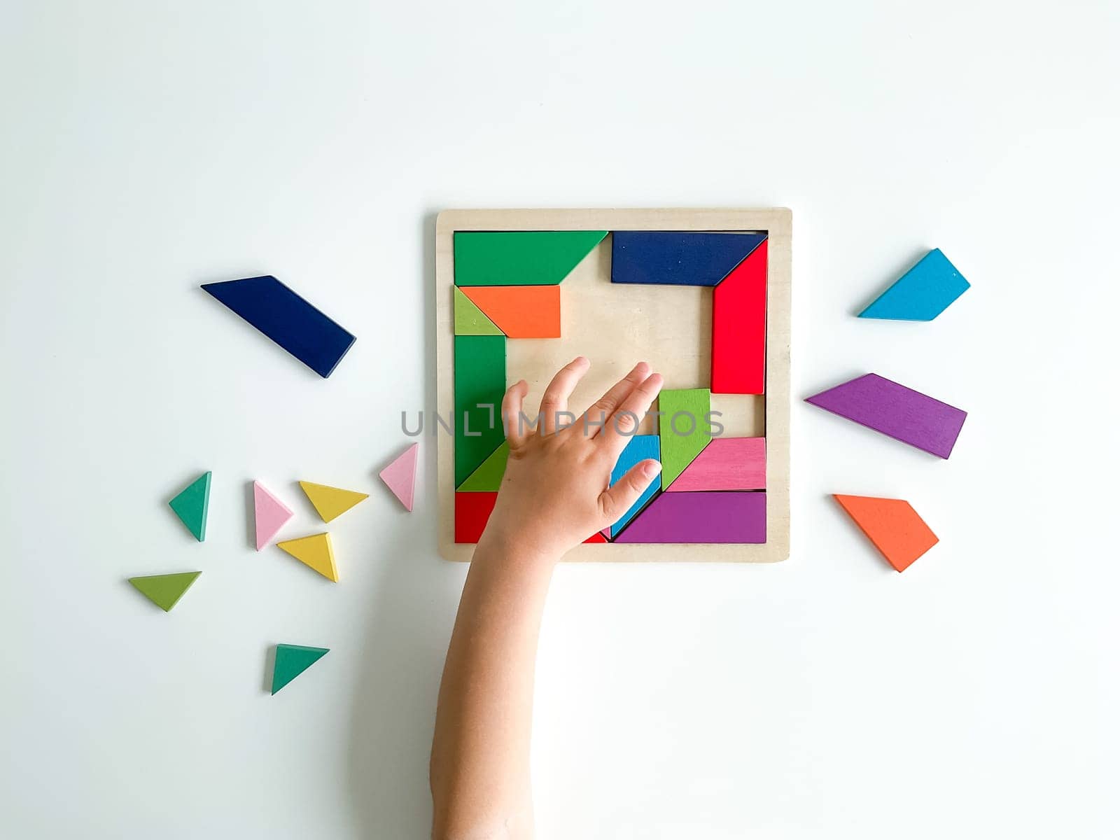 childs hand collects multicolored wooden mosaic on white background. child solves a colorful tangram. square of colorful geometric shapes on white background.