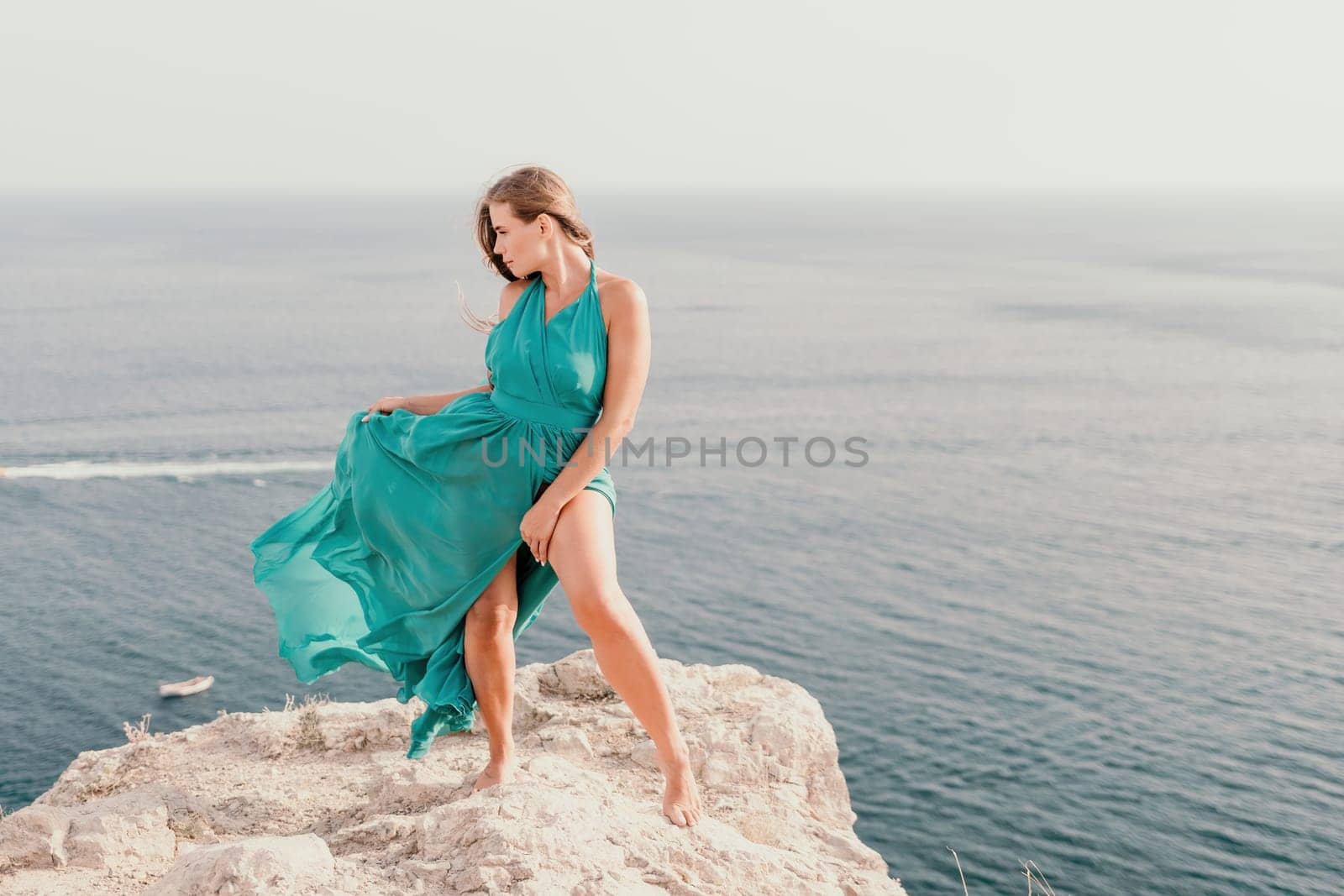 Side view a Young beautiful sensual woman in a mint long dress posing on a volcanic rock high above the sea during sunset. Girl on the nature on overcast sky background. Fashion photo