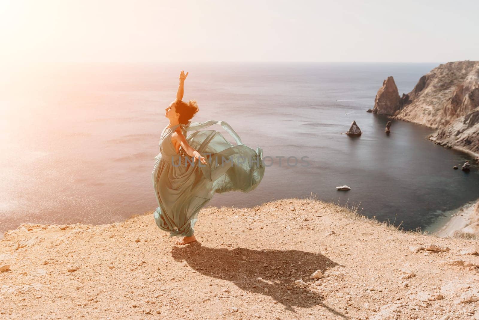 Side view a Young beautiful sensual woman in a red long dress posing on a rock high above the sea during sunrise. Girl on the nature on blue sky background. Fashion photo.