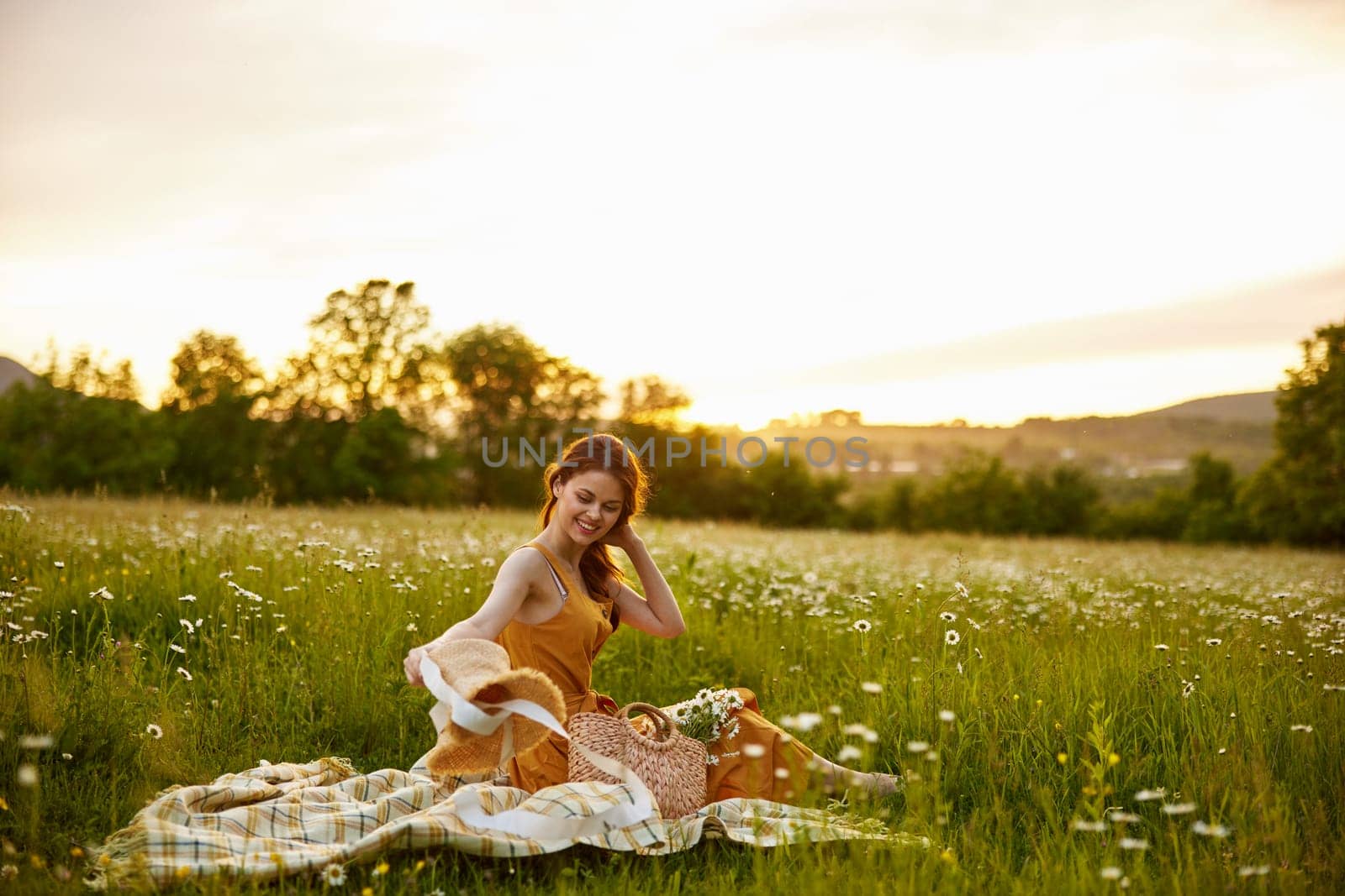 a happy woman in an orange dress sits in a meadow of daisies during sunset and looks at her hat. High quality photo