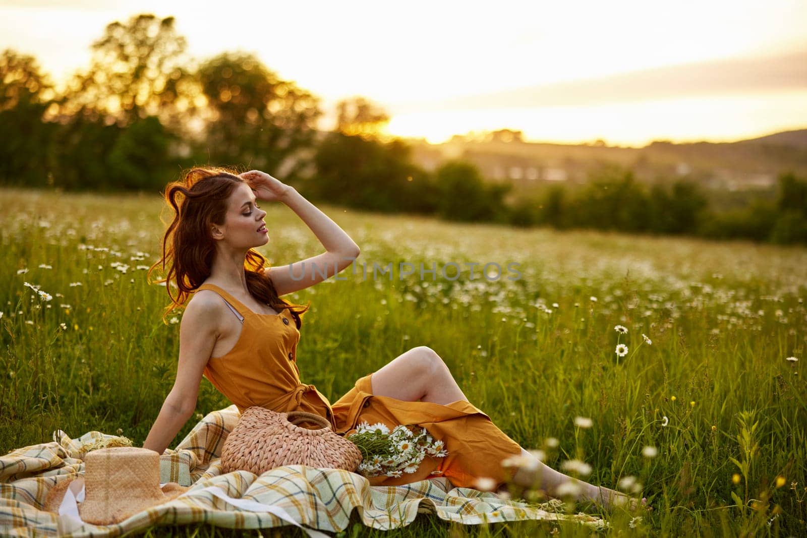 a beautiful woman in an orange dress sits on a plaid in a chamomile field and straightens her hair. High quality photo