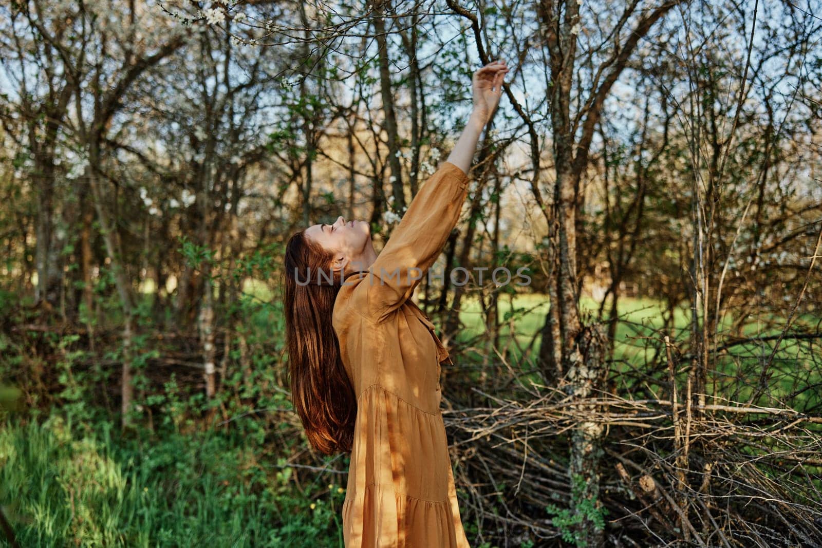 a beautiful, slender woman with long hair walks in the shade near the trees, dressed in a long orange dress, enjoying the weather and the weekend, joyfully raising her hands up. High quality photo
