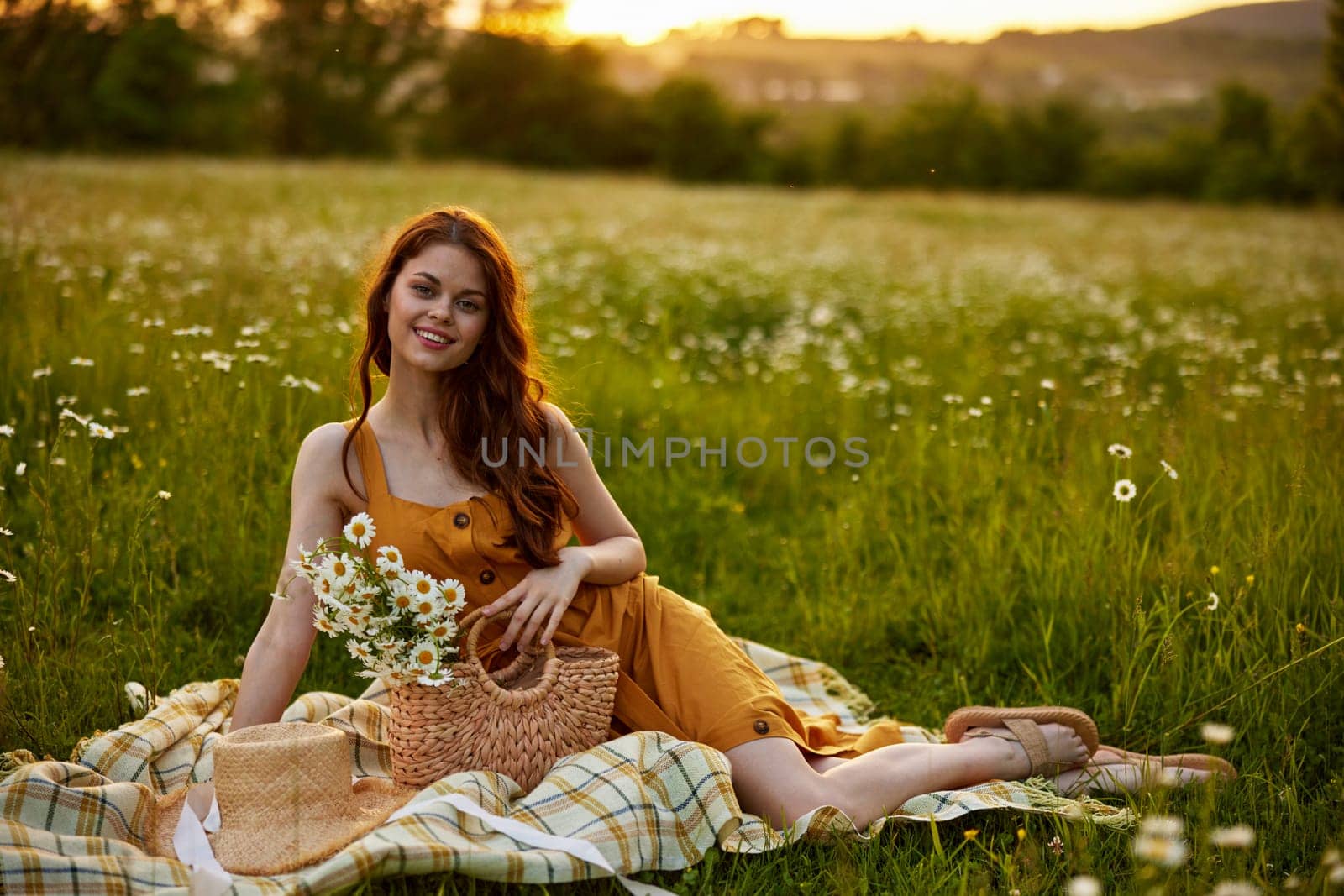 a happy woman in an orange dress sits on a plaid in a chamomile field at sunset and laughs. High quality photo