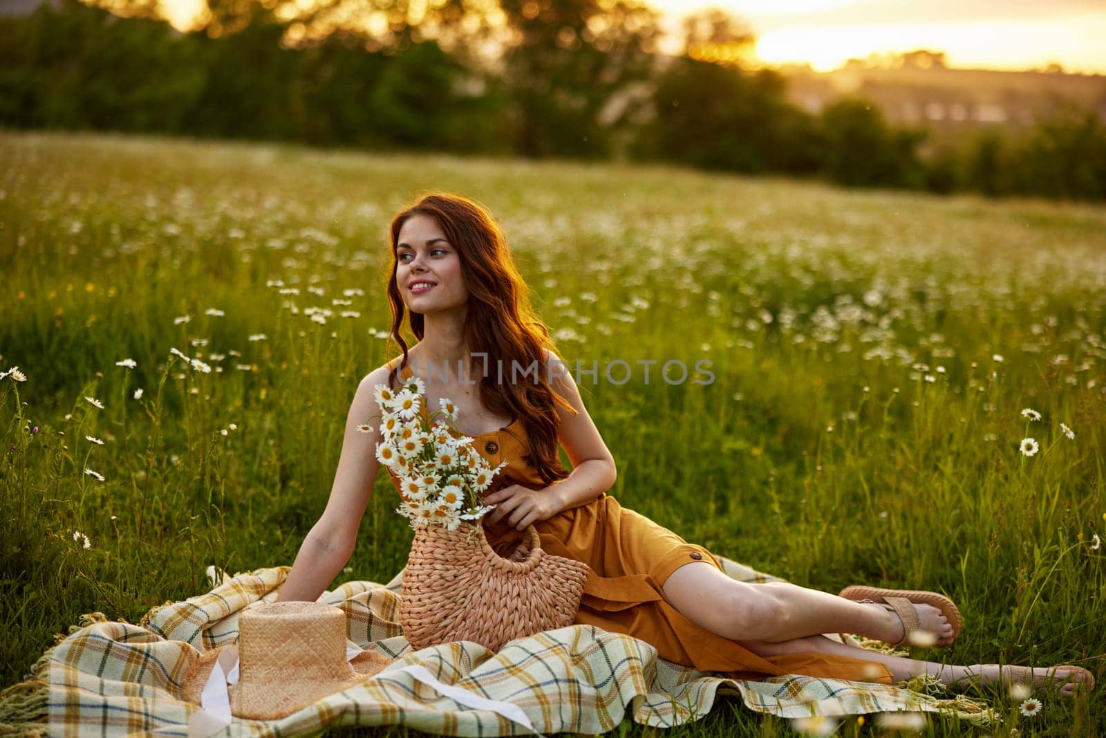a happy woman in an orange dress sits on a plaid in a chamomile field at sunset and laughs by Vichizh