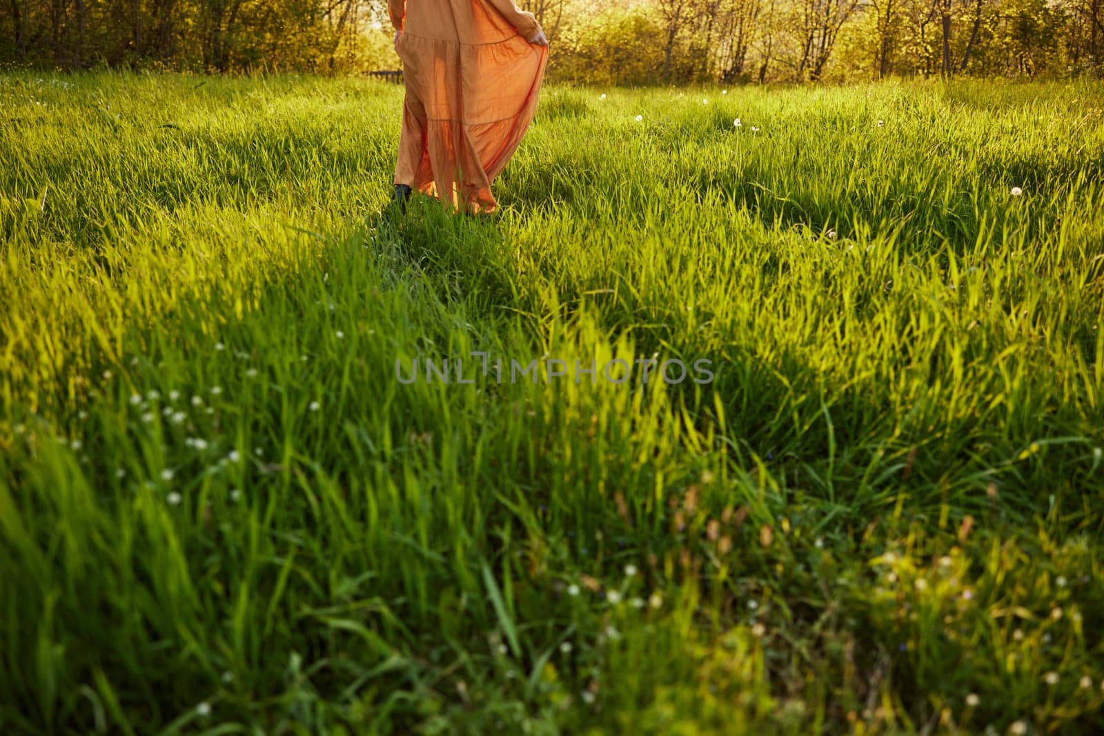horizontal photo of a woman in an orange dress photographed without a face, standing in a green field during sunset, illuminated from the back by Vichizh