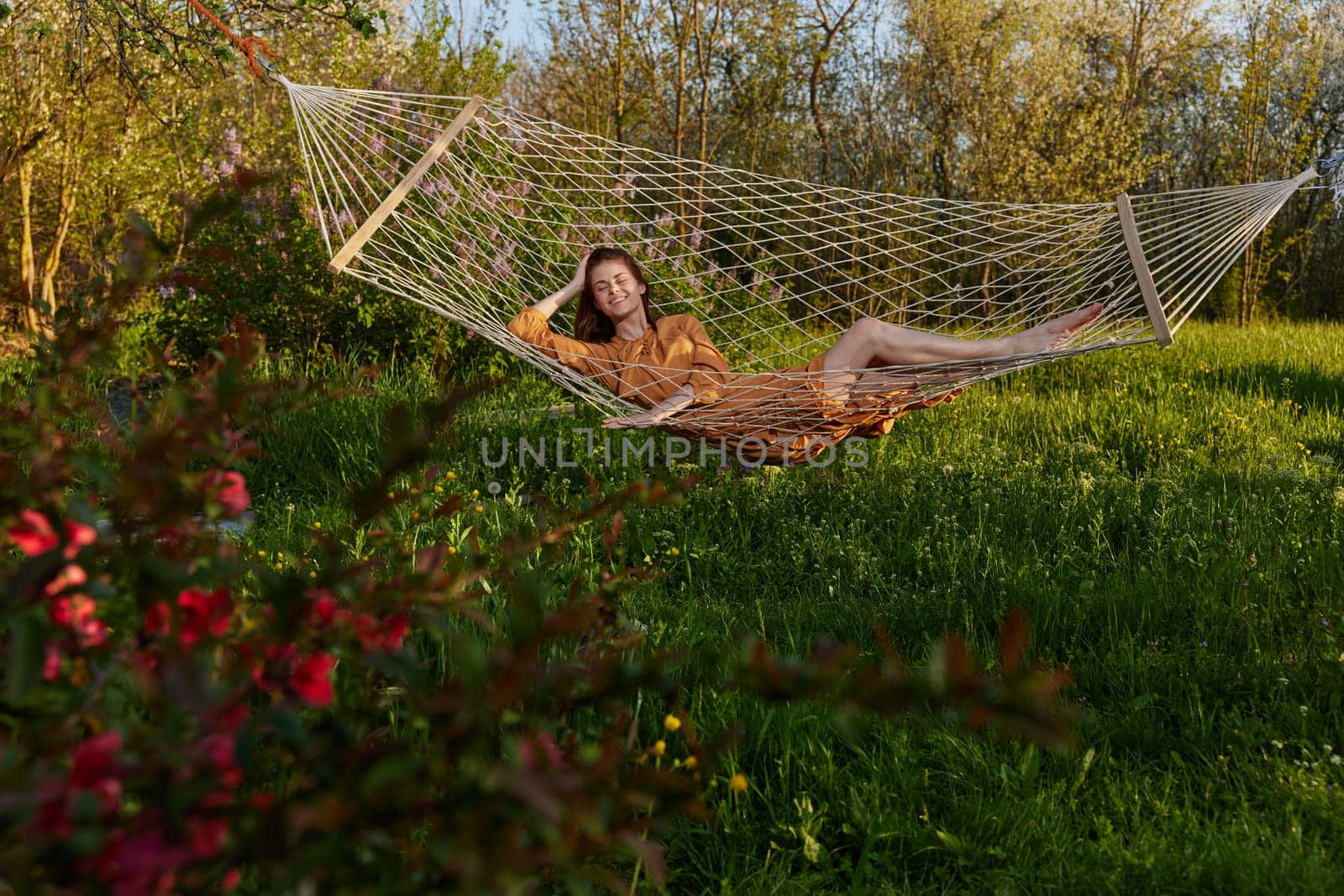 a happy woman in a long orange dress is relaxing in nature lying in a mesh hammock enjoying summer and vacation, looking at the sky. High quality photo