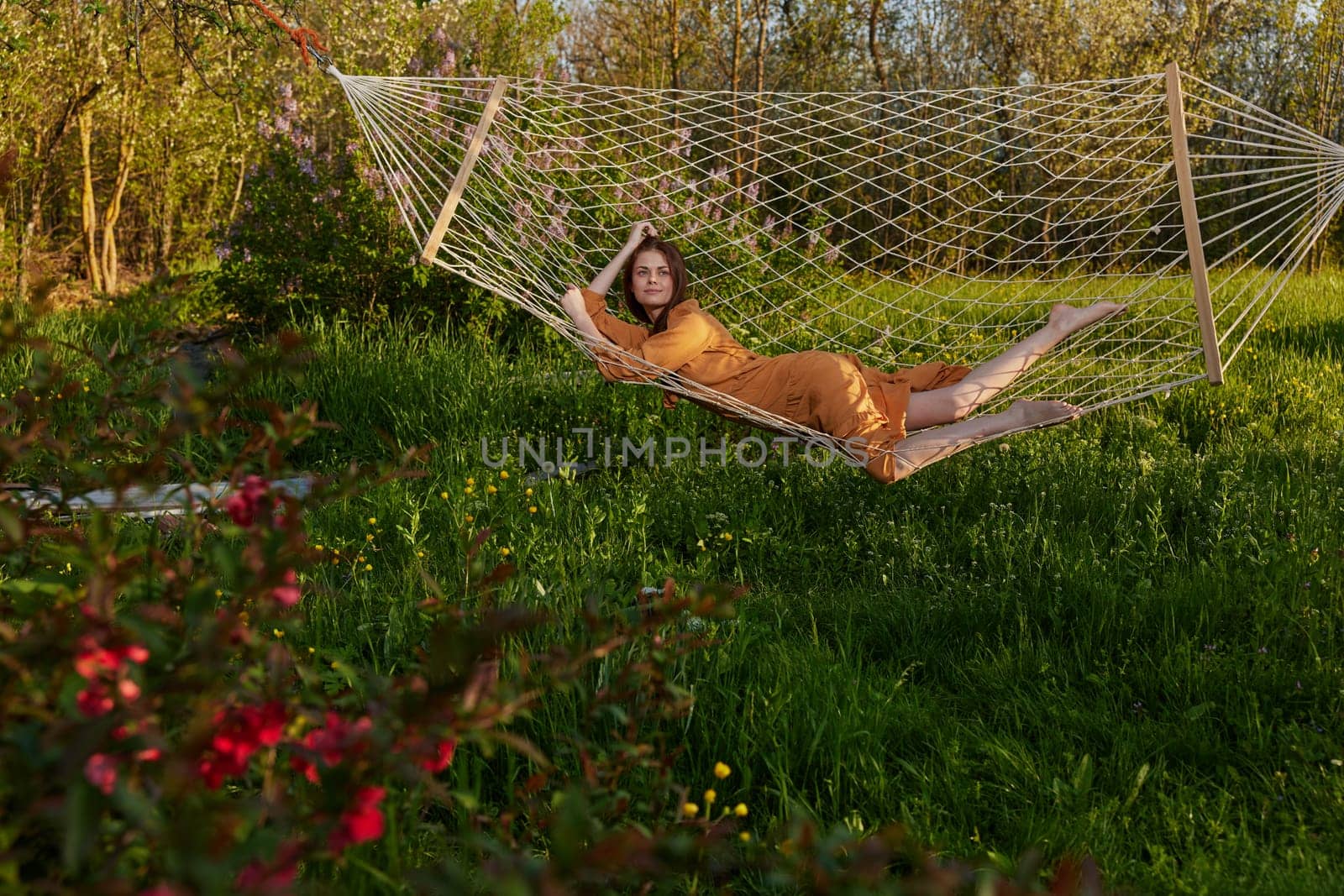a happy woman in a long orange dress is relaxing in nature lying in a mesh hammock enjoying summer and vacation in the country surrounded by green foliage. High quality photo