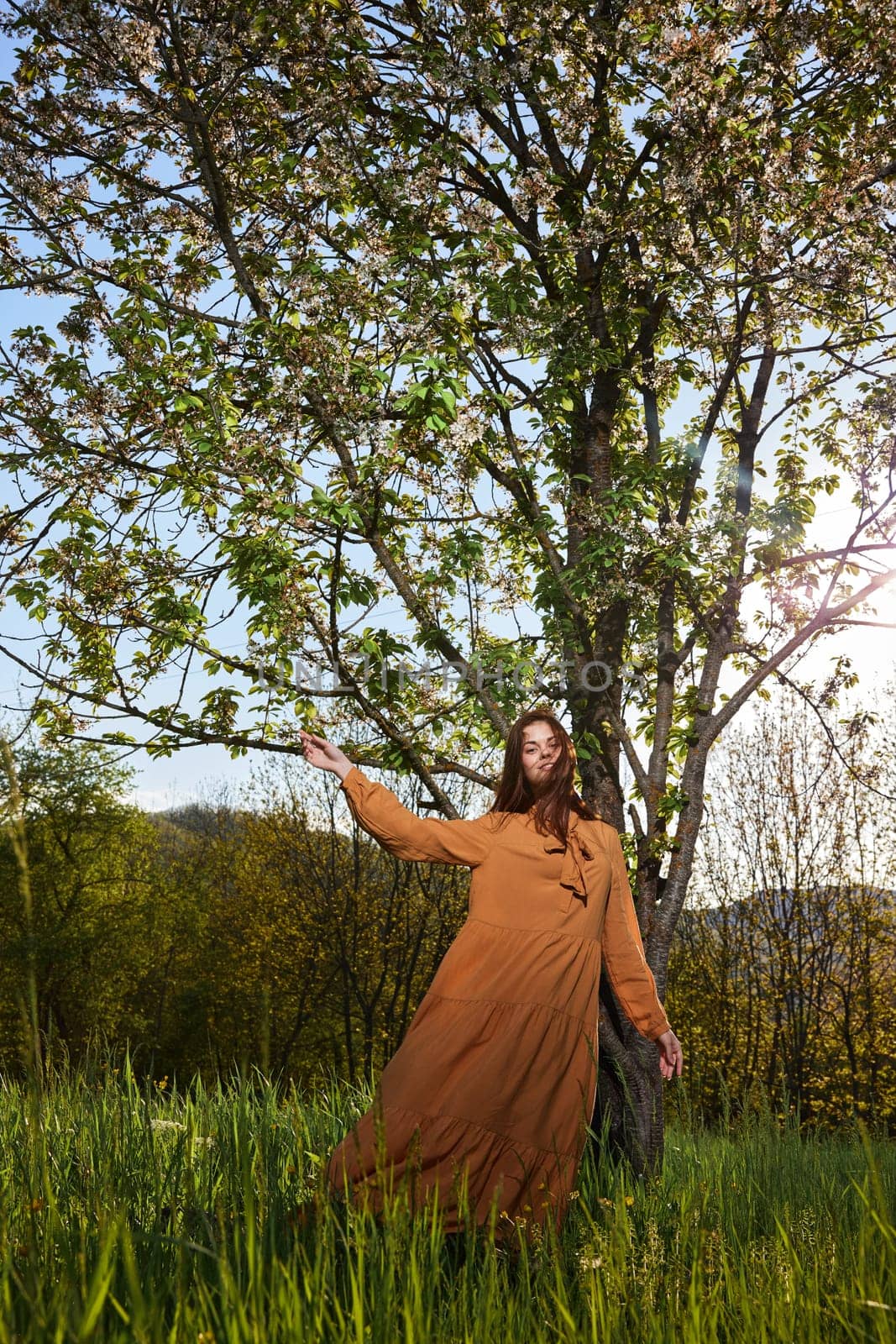 a slender, attractive woman with long red hair is standing in the countryside near a flowering tree in a long orange dress and happily spinning holding the dress. Vertical photography. High quality photo