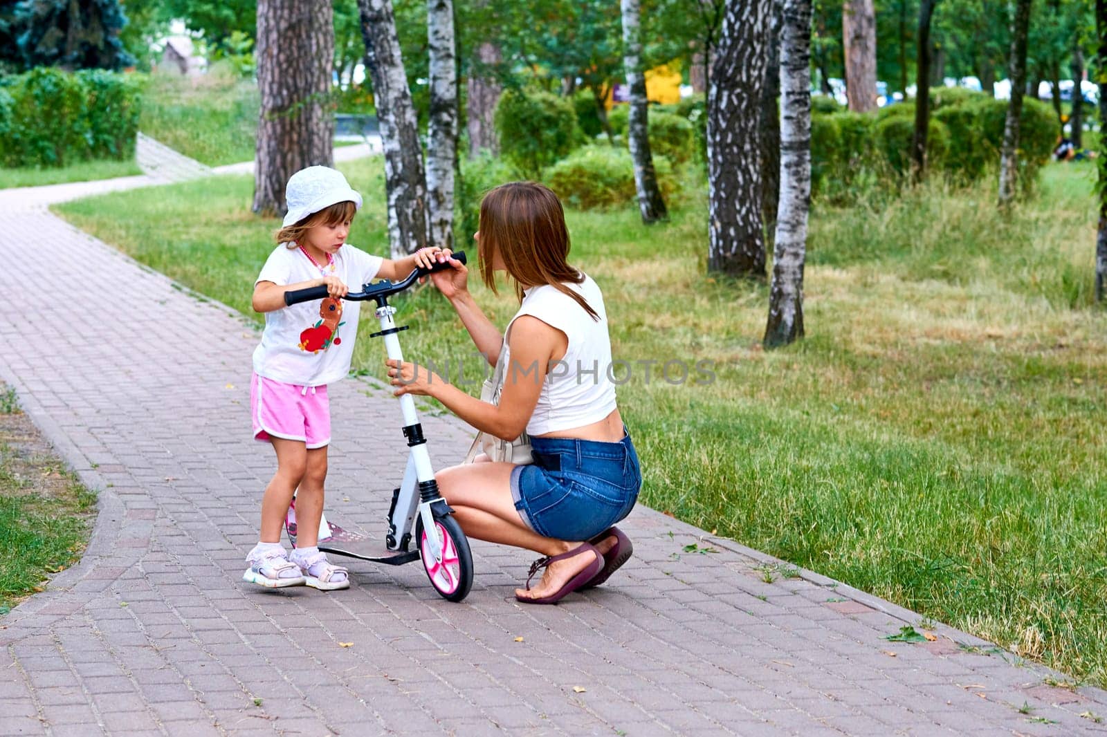 A young mother raises educates a child with a scooter on a path in a summer park by jovani68