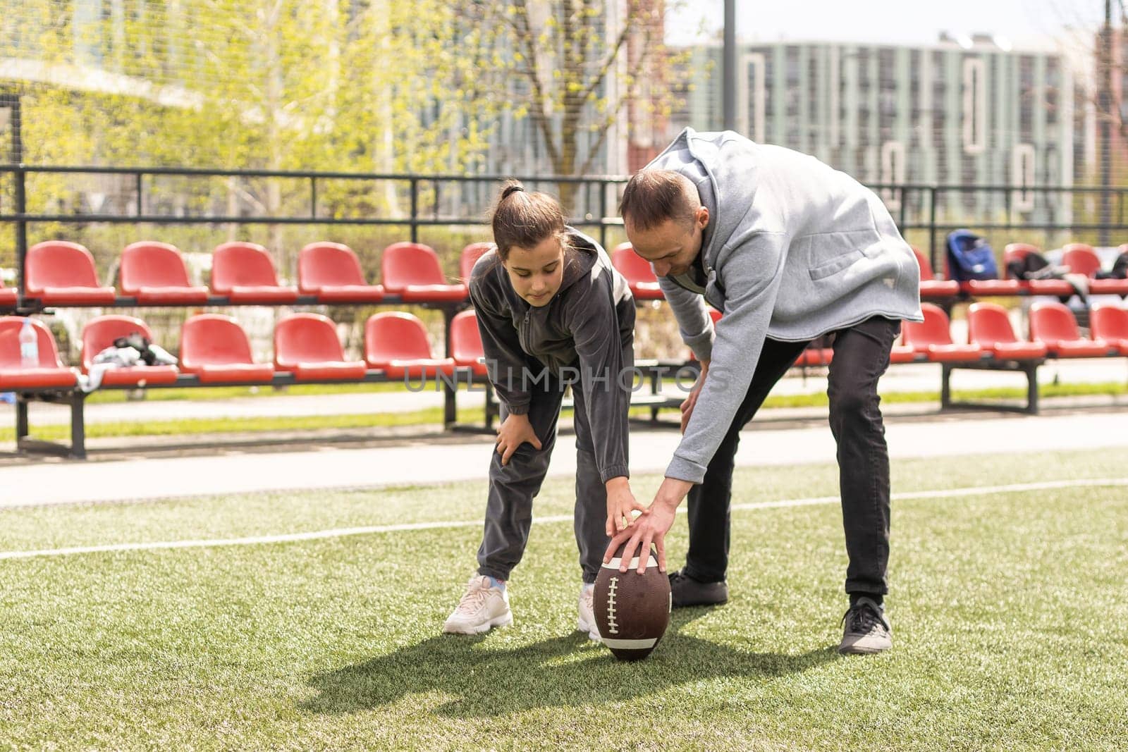 Handsome dad with his little cute daughter are having fun and playing American football on green grassy lawn.