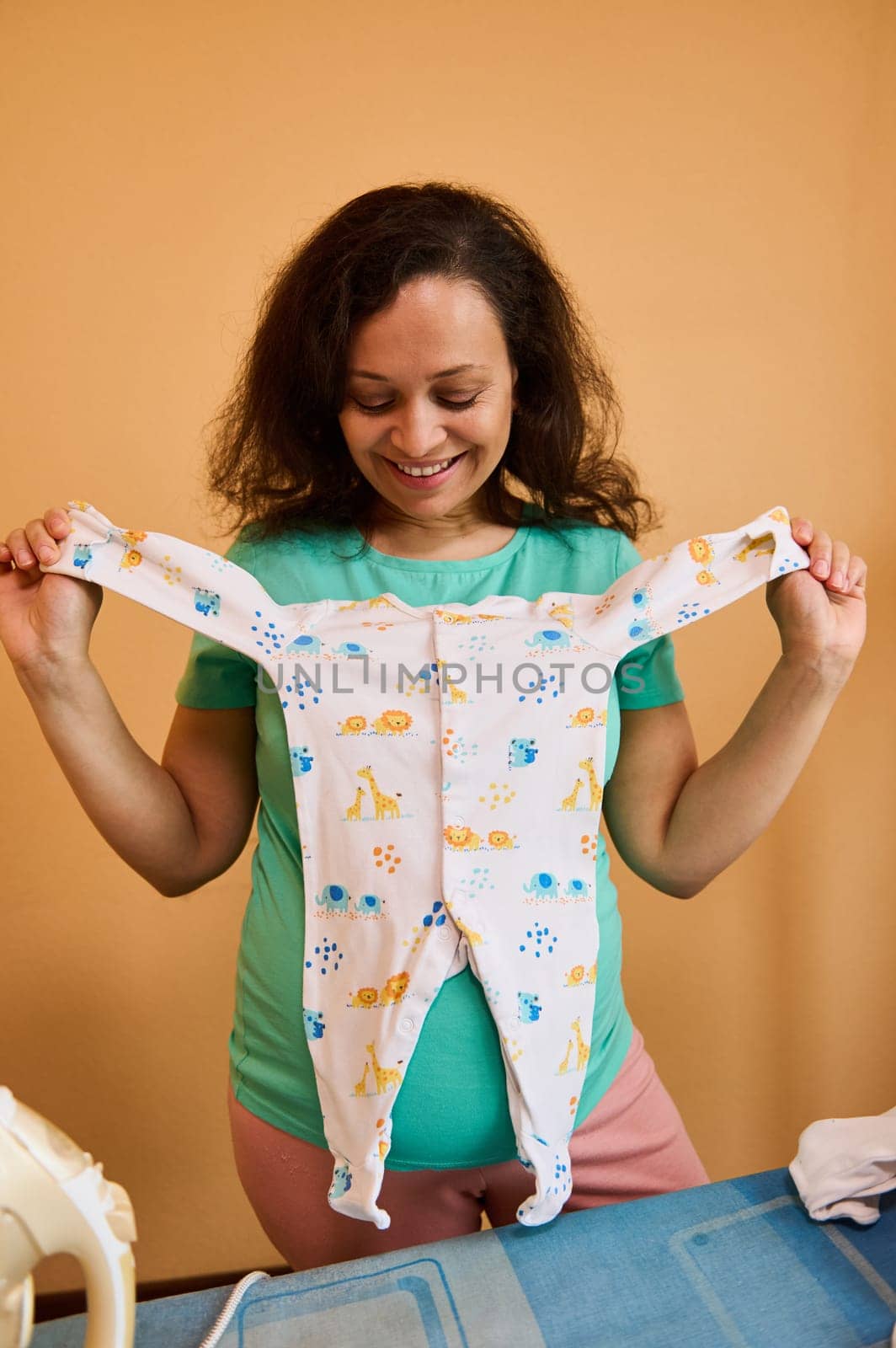 Happy smiling pregnant woman holding a cute jumpsuit for her newborn baby, standing by ironing desk while preparing bag for maternity hospital. Motherhood. Pregnancy last trimester. People. Lifestyle.