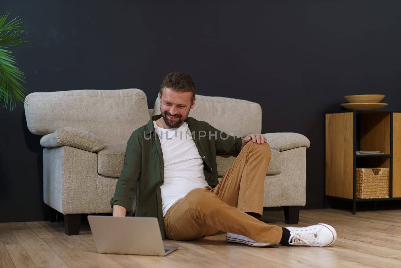 Man sitting on floor near sofa in house and working on laptop. He is freelancer or remote worker, using technology and internet to do job. by LipikStockMedia
