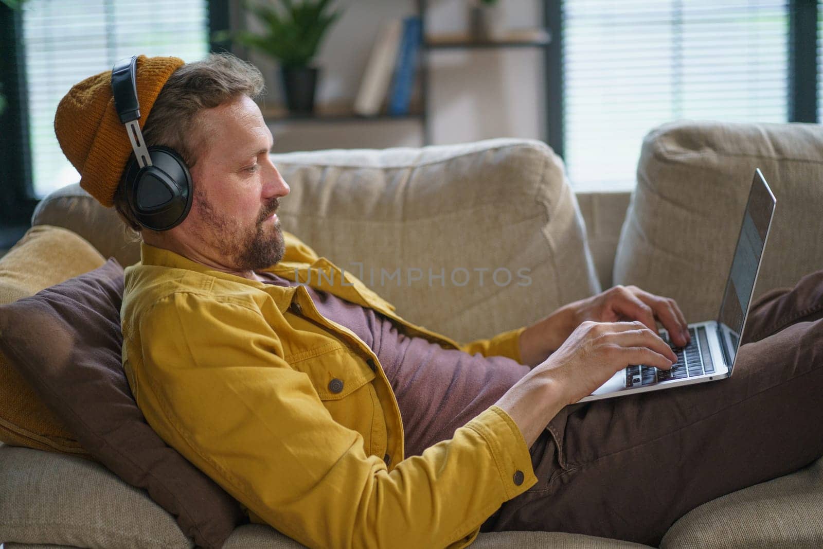 Young man in casual clothing sitting on sofa home, playing online game on laptop. He is fully concentrated on game, wearing headphones and using the keyboard to control action on screen. High quality photo