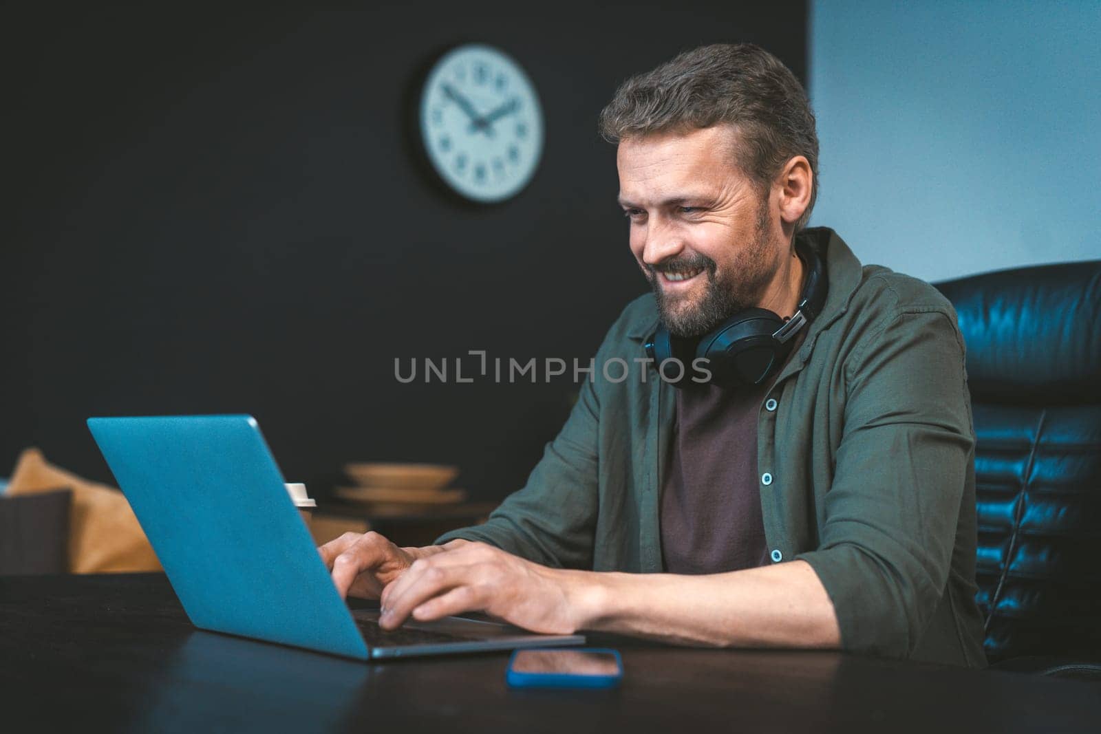 Happy and motivated man is working on laptop at desk in stylish loft office, with intention of making money. He focused and determined, yet also relaxed and comfortable in work environment. High quality photo