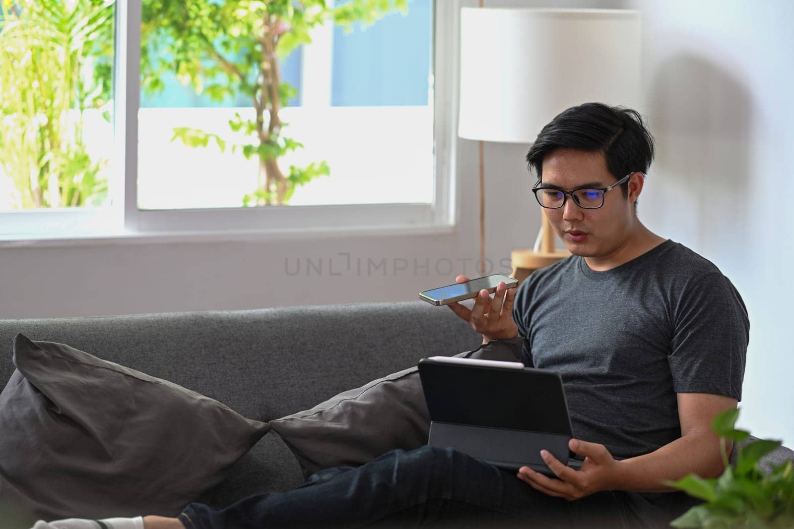 Handsome man talking on speaker phone and using computer tablet on sofa.