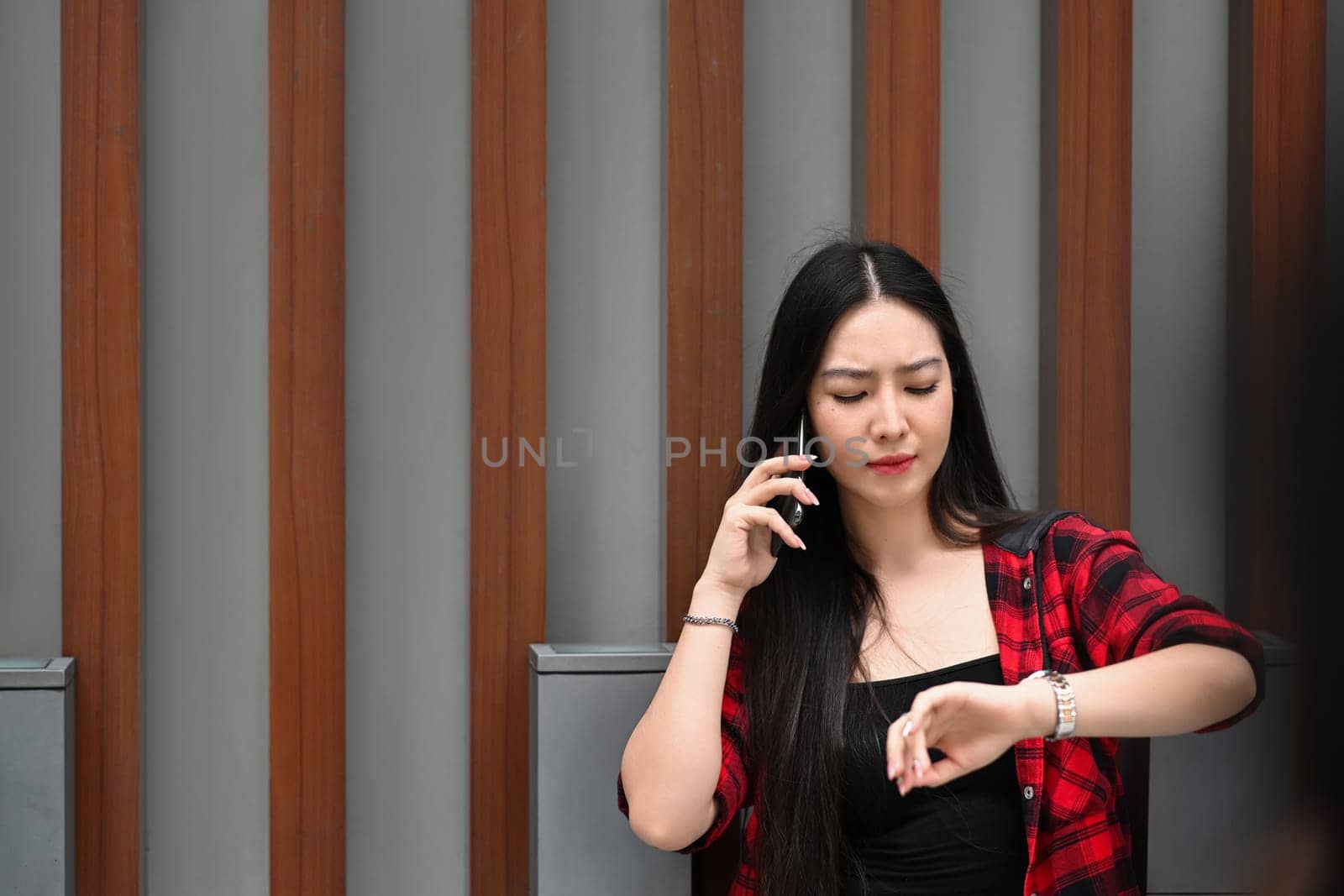 Young woman talking on mobile phone and checking time on her wrist watch while standing outdoor in the city. by prathanchorruangsak