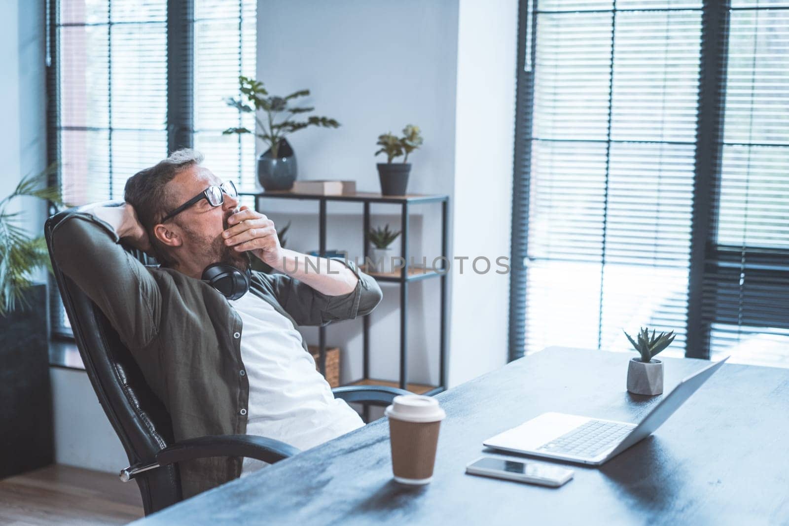 Tired and overworked office worker sitting at desk in office, working on notebook. Worker appears exhausted and bored, likely due stress and fatigue of meeting work deadlines and multitasking. High quality photo