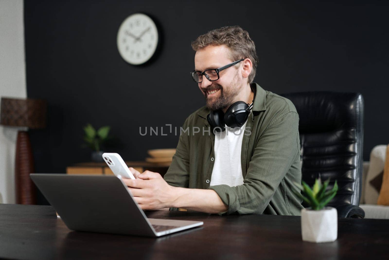 Concept of communication and technology in workplace. European man texting on mobile phone while working on laptop in office. Multitasking, concentration, and seamless connection between two devices. High quality photo
