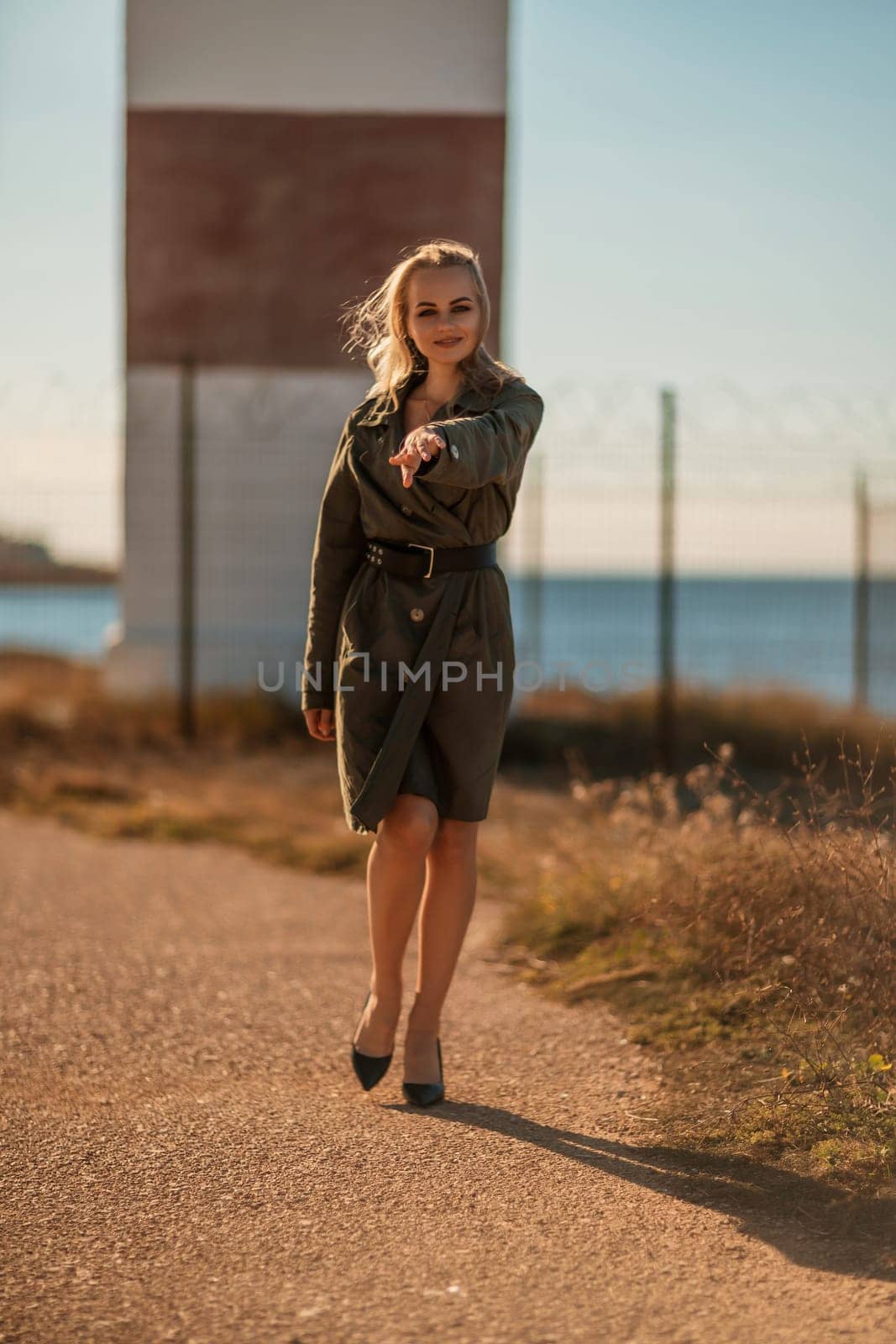 Portrait blonde sea cape. A calm young blonde in a khaki raincoat stands on the seashore against the backdrop of a lighthouse