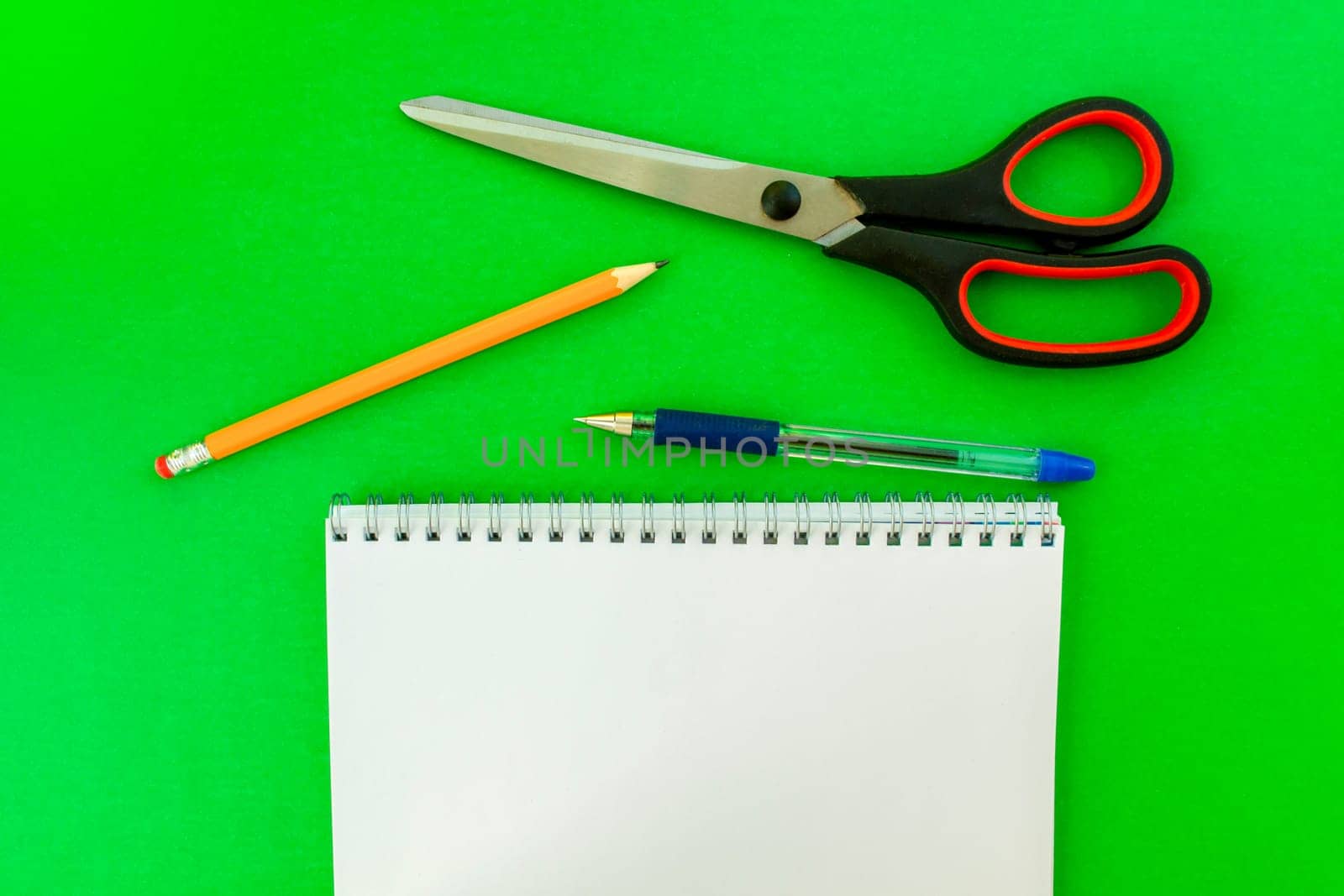 White notebook, pen, pencil and scissors. School white notepad with spring, pen, pencil and scissors on a green background.
