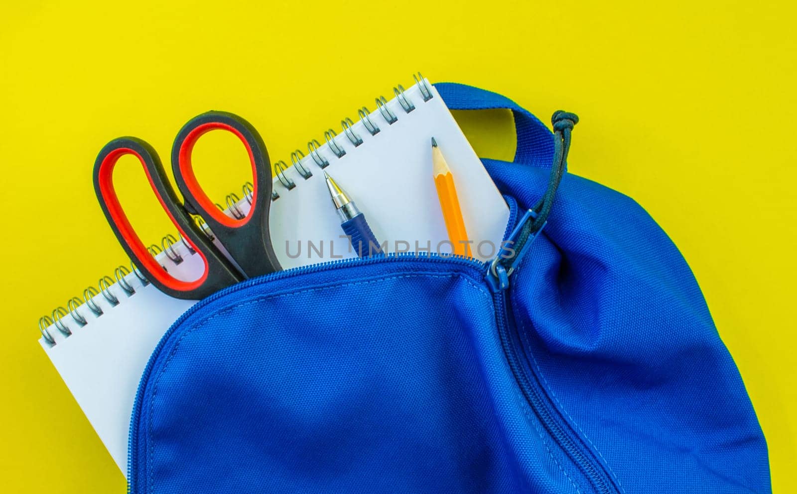 School backpack, pencil, pen, scissors and notepad on a yellow background. School blue backpack with notepad, pencil, scissors and pen inside on a yellow background.