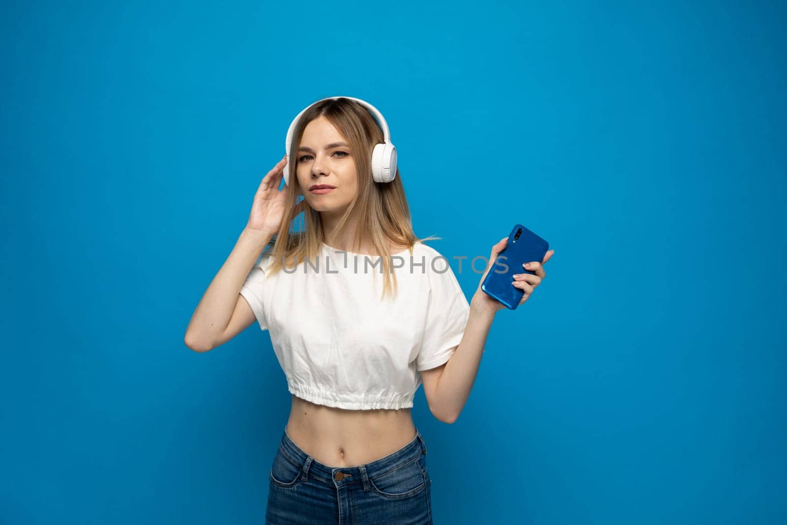 Young blonde woman in white t-shirt with headphones listening music. Music teenager girl dancing against isolated blue background