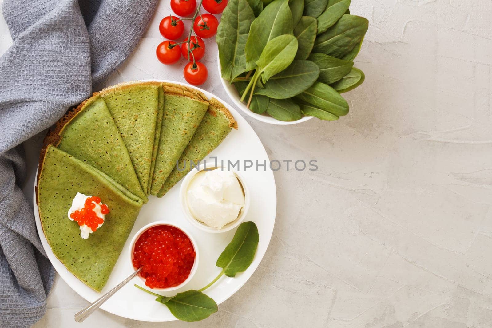 Spinach pancakes on a plate with soft cheese, caviar and cherry tomatoes on a gray background with textile. copy space