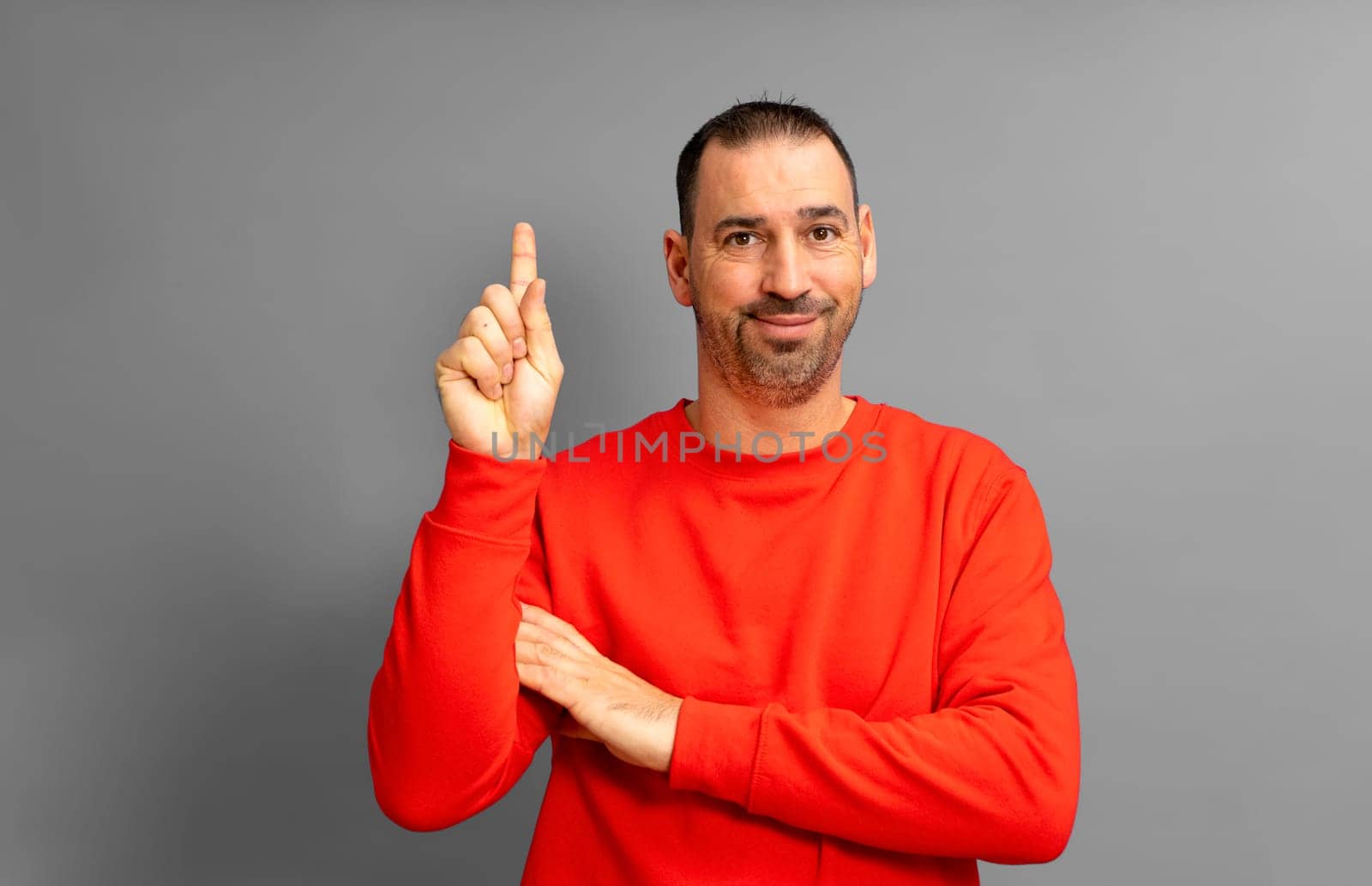 Proactive smart insightful hispanic man 40s wearing red jumper holding index finger up with great new idea isolated on gray background studio portrait. People lifestyle concept.