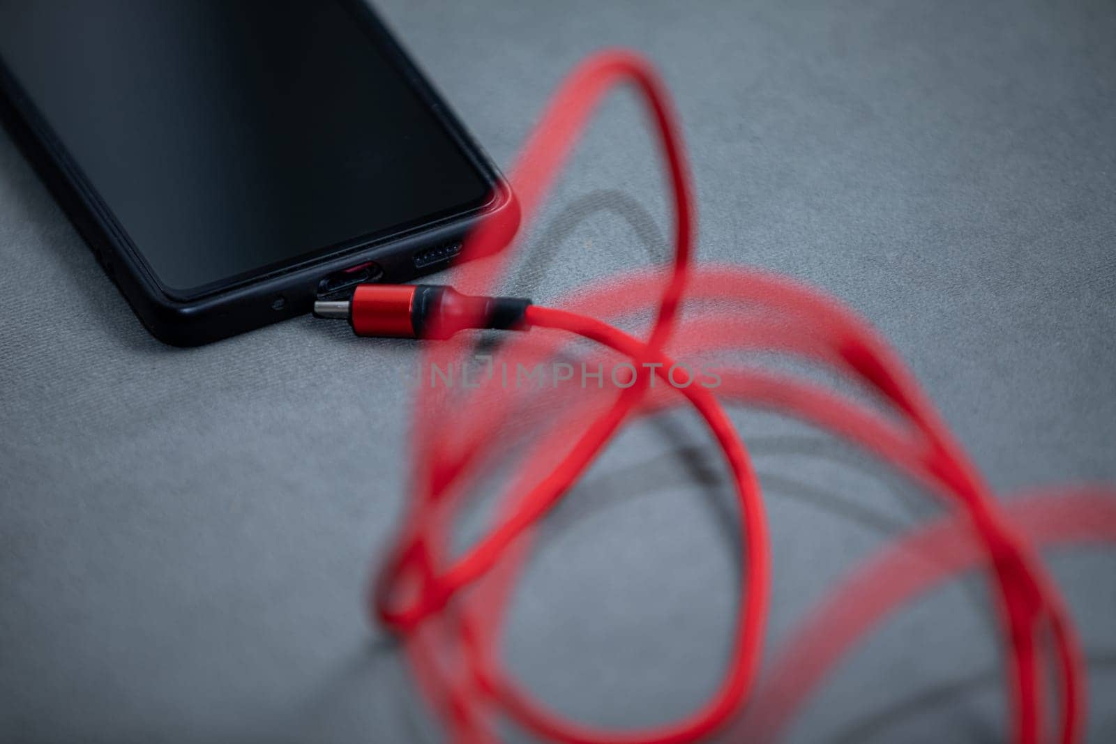 On the gray fabric lies a black smartphone and and red charger cable. Smartphones need to be charged frequently.