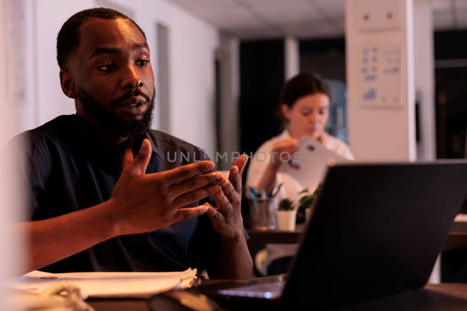 Project manager discussing startup with colleague on videoconference, corporate worker answering internet call in office at night. African american man using online meeting computer app