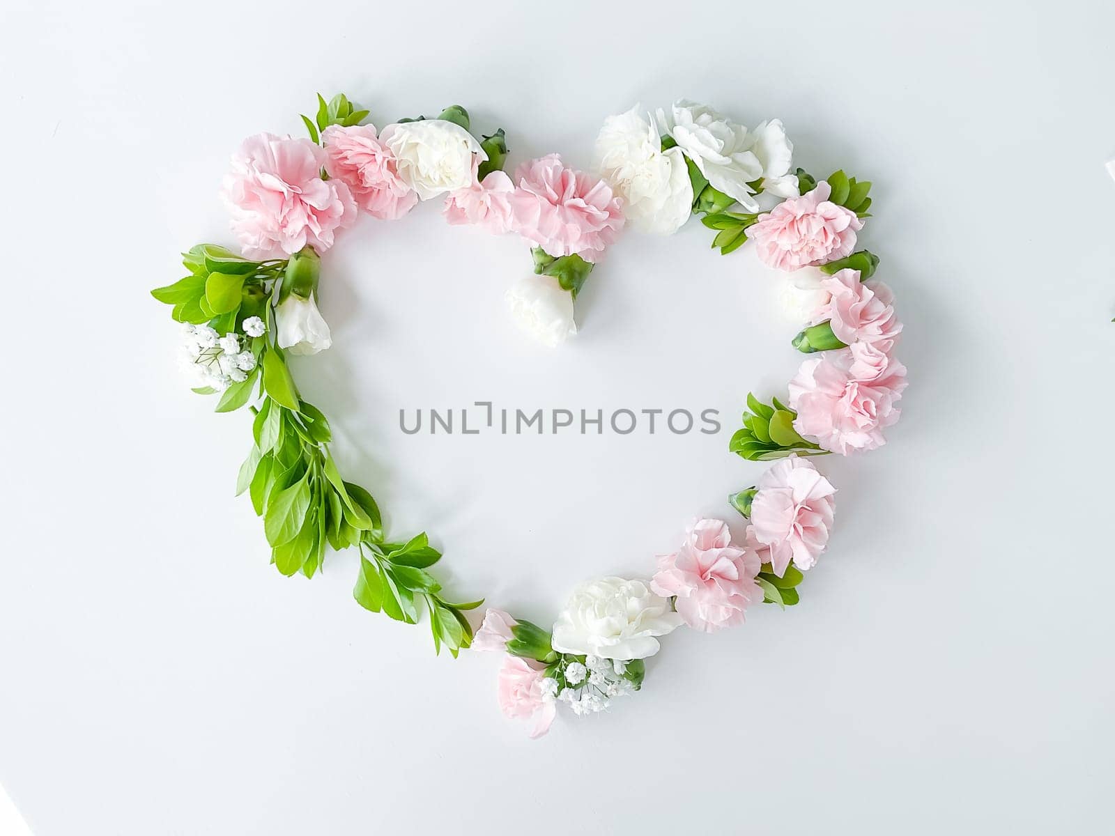 Frame in the form of a heart from pink and white carnations, green leaves, gypsophila on a white background. Flat lay, top view. Spring background. Suitable for wedding, cards and invitations