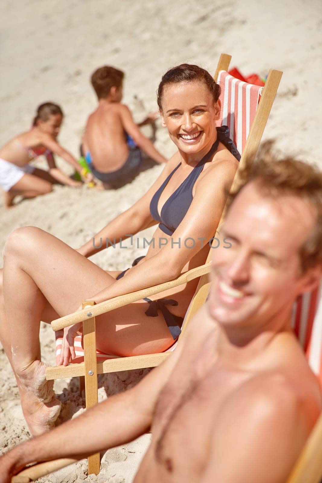 Soaking up some summer sun. a couple sitting on beach chairs while their children play in the background. by YuriArcurs