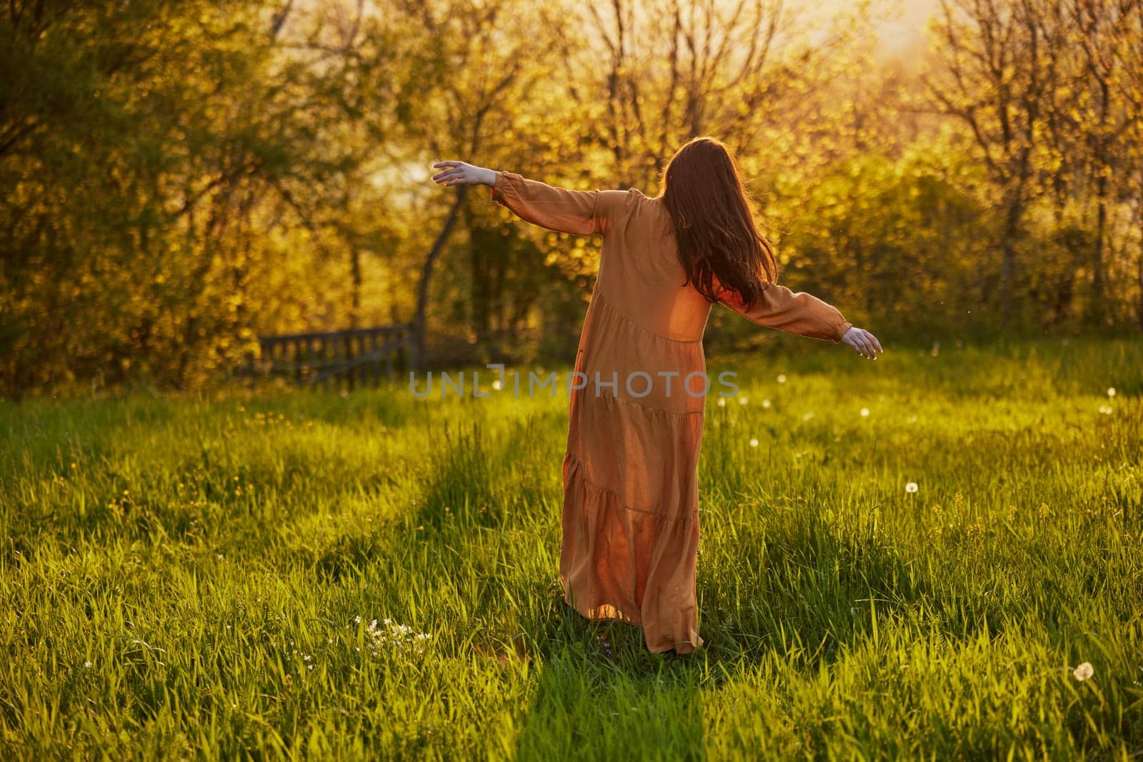 a slender woman with long hair stands in a field with her back to the camera, illuminated by the rays of the setting sun and happily poses enjoying the warm weather and rest. High quality photo